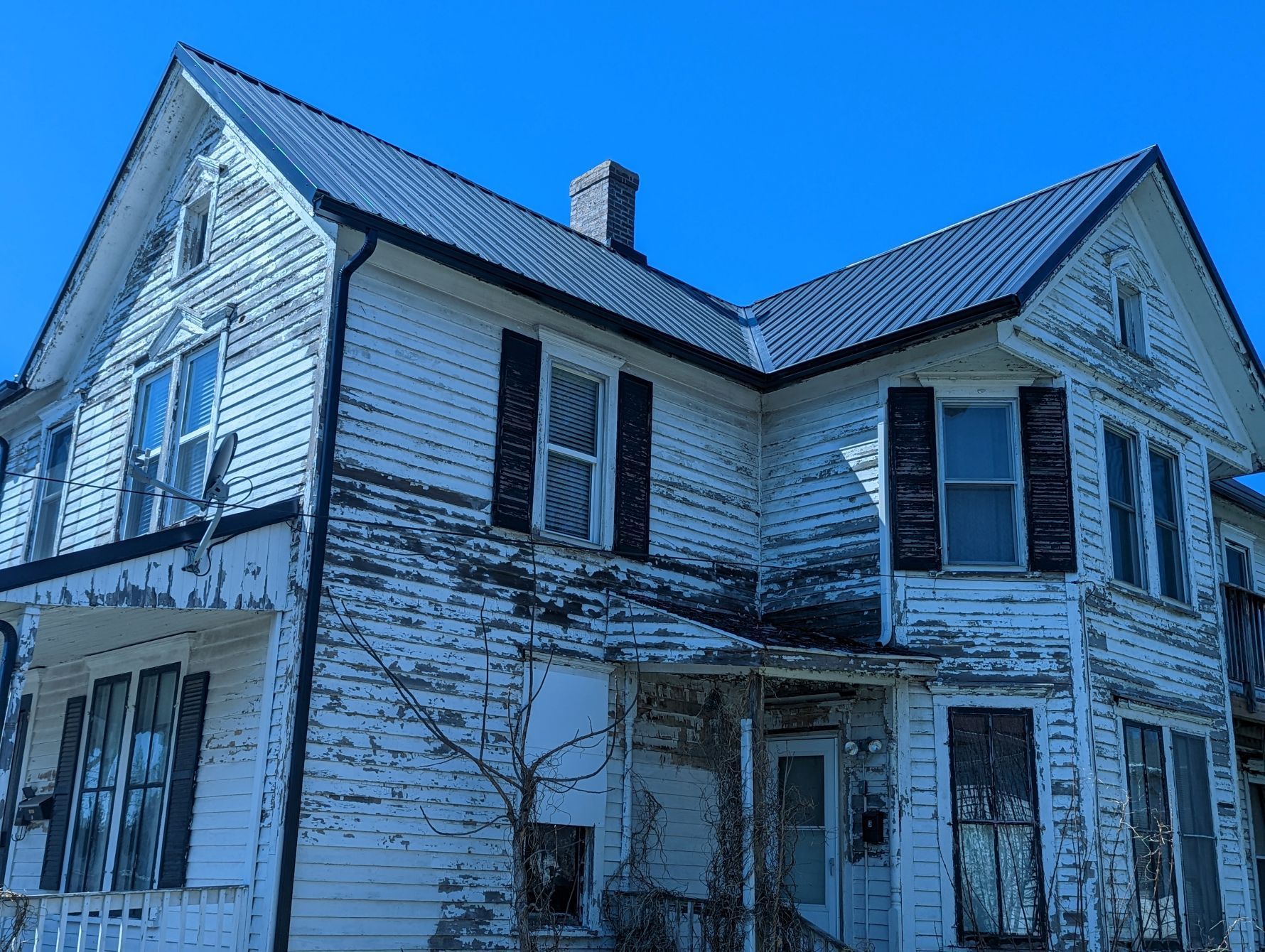 Old white house with peeling paint, black shutters, and metal roof against a blue sky.