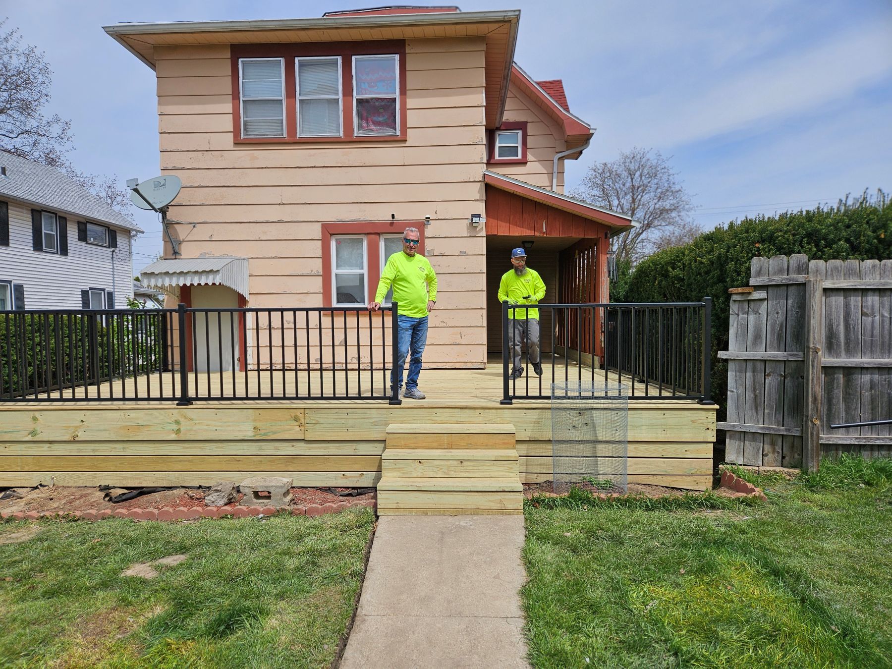 Two people stand on a newly constructed wooden deck in front of a house. Black railing surrounds the deck.