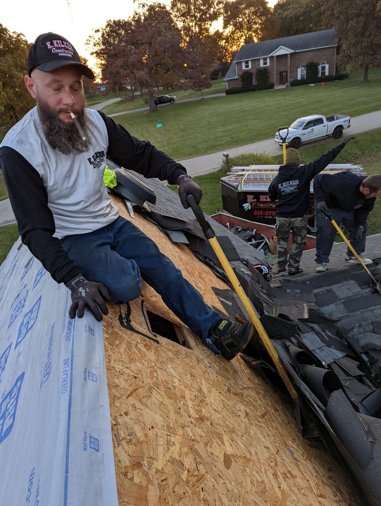 Man on roof with a beard smoking while removing shingles with tools; two other workers are in the background.