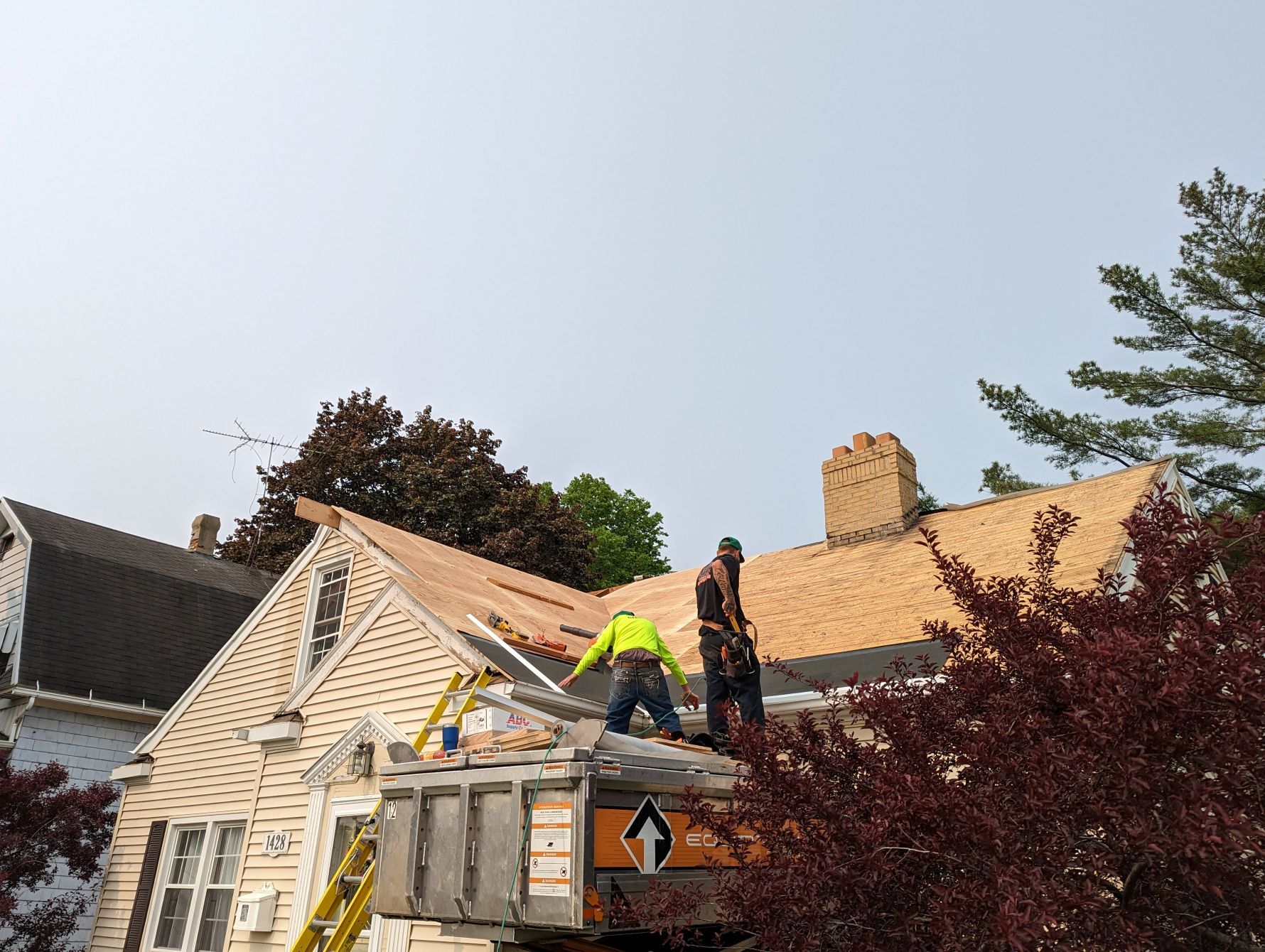 Two roofers working on a house roof. One wears a yellow vest, other in black. Sky visible.