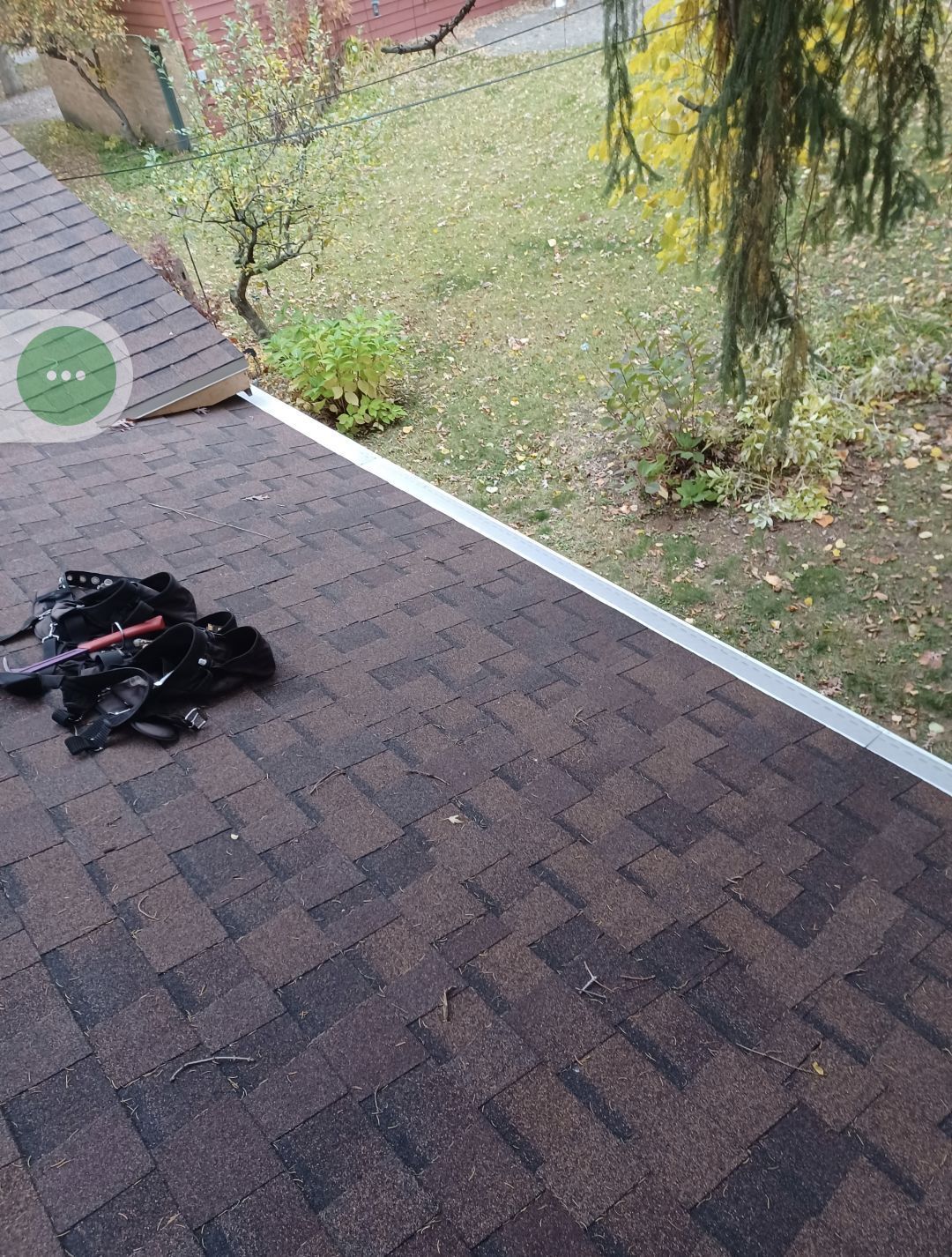 Roof with brown shingles, silver gutter. Black tools on shingles, autumn yard in background.