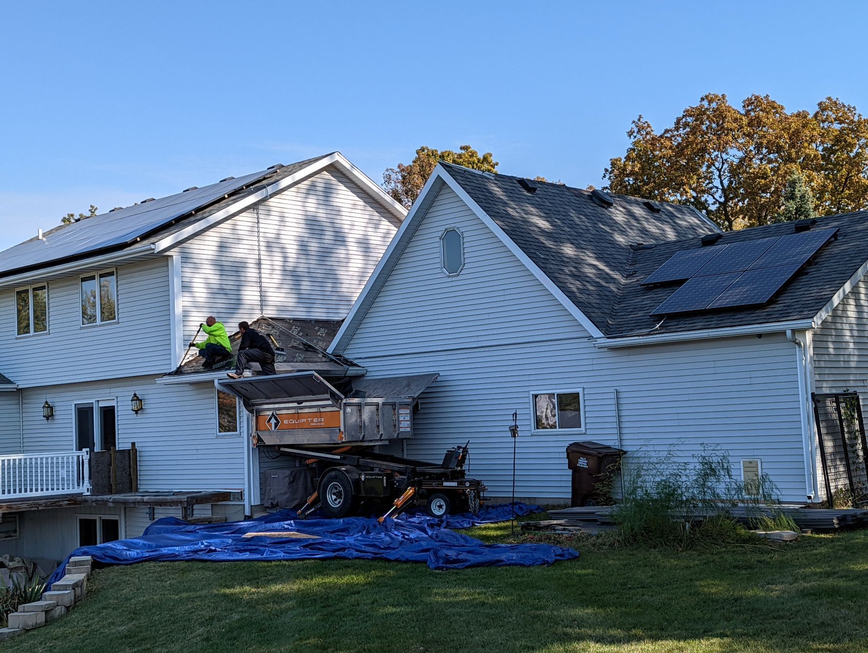 Roofers on a house with solar panels, using a debris removal system; blue tarp on lawn, sunny day.