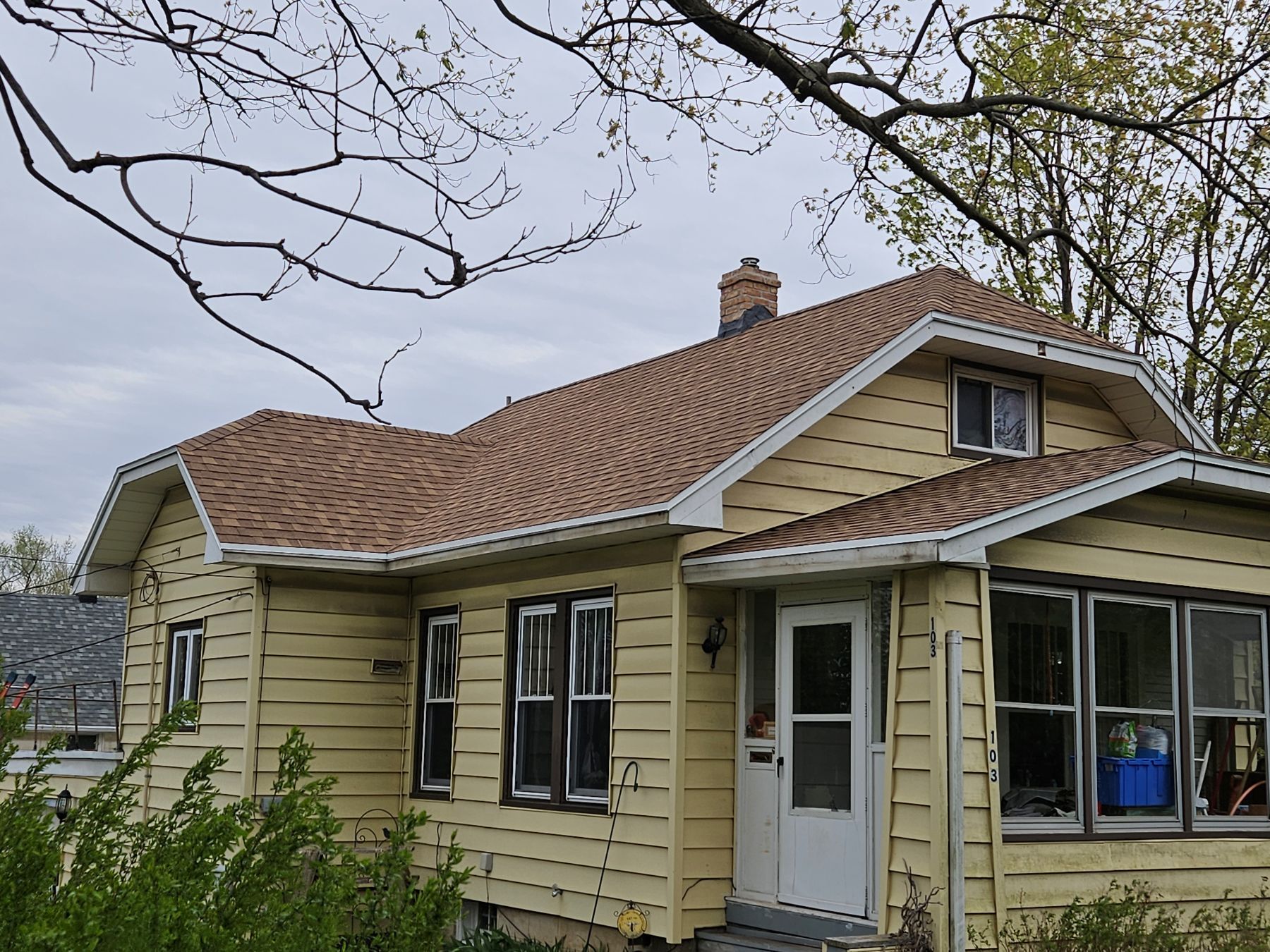Yellow house with brown roof, windows, and white door under a cloudy sky.