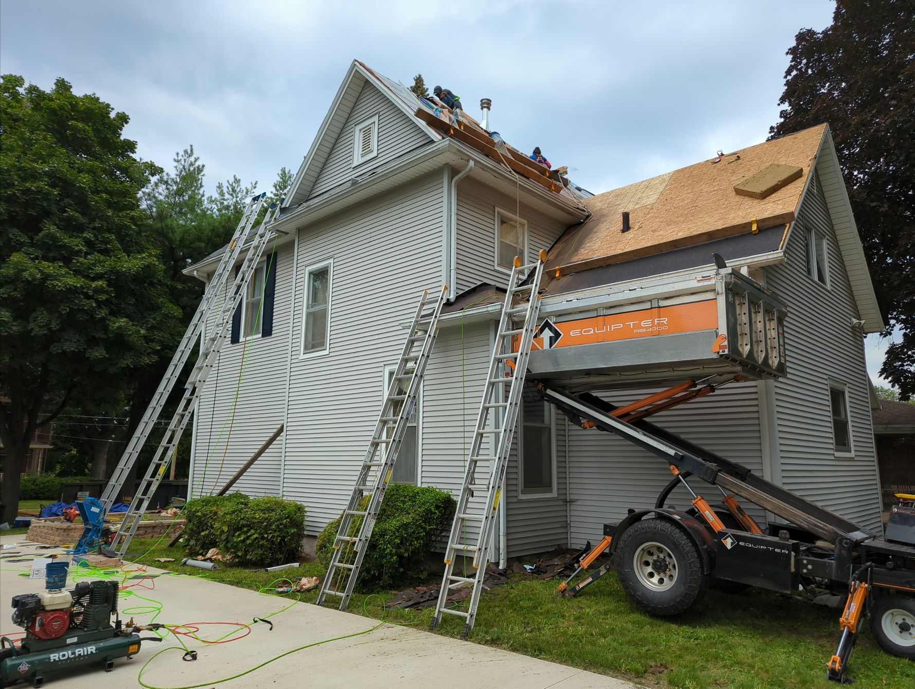 House under construction, roof being replaced. Workers on roof and ladders, lift equipment on grass. Overcast sky.