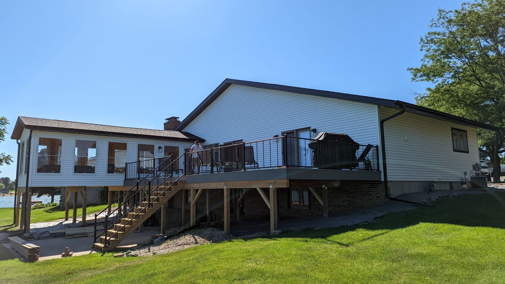 House with a large deck and enclosed porch overlooking a grassy area and body of water under a blue sky.