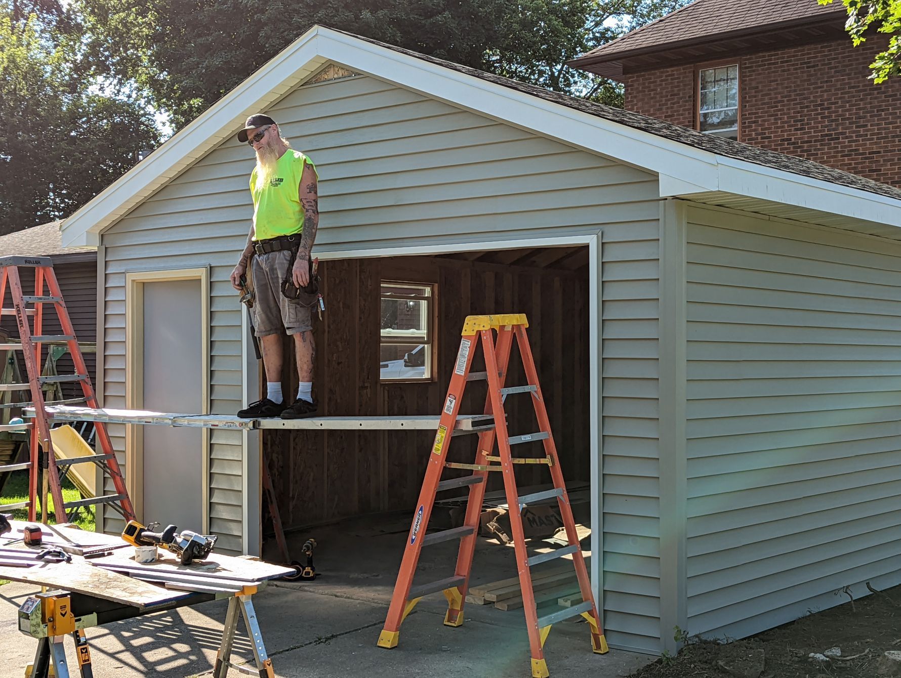 Man standing on a platform, working on a garage. Exterior shot, light siding, orange ladder.