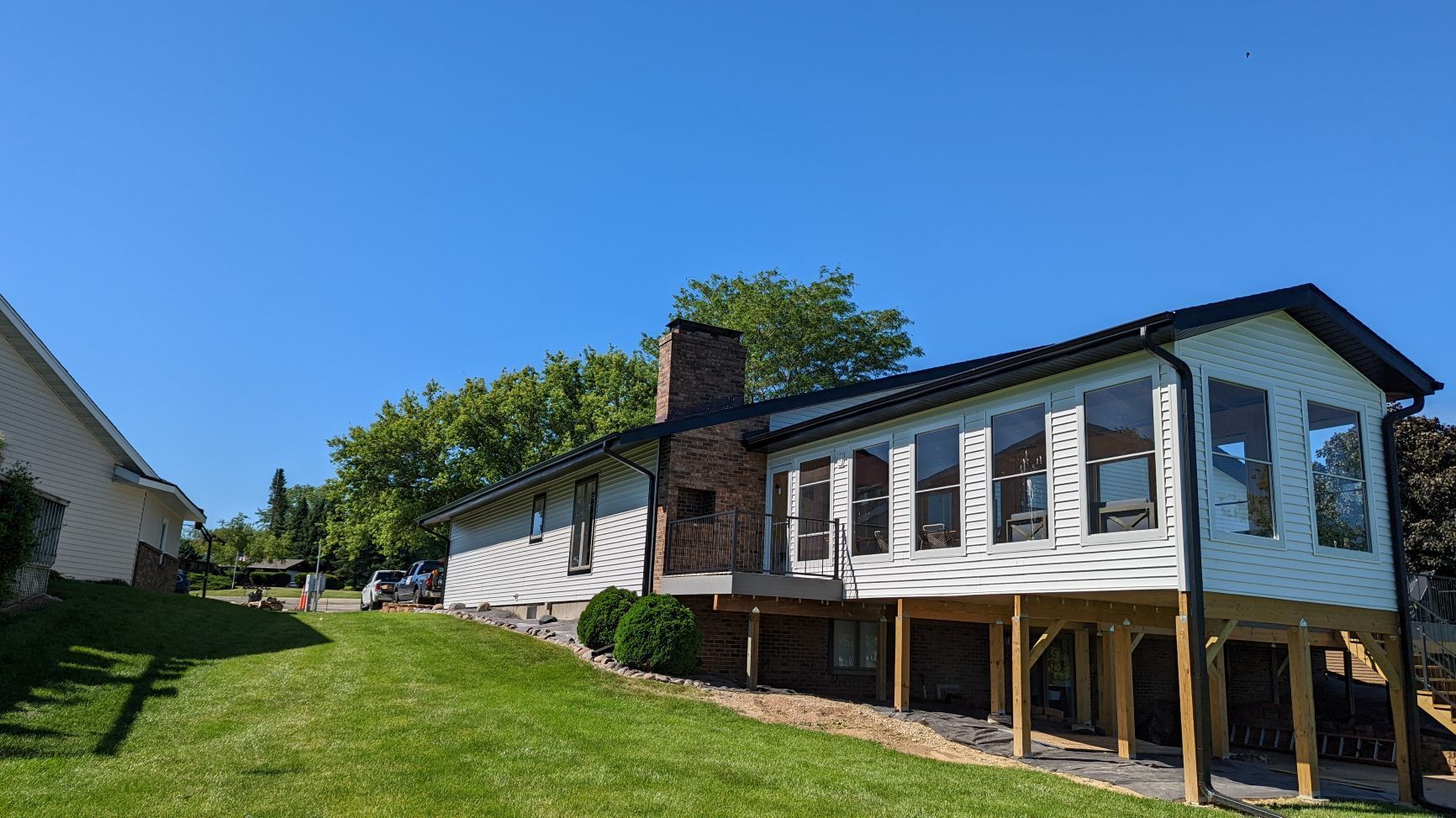Side view of a house with white siding, wooden deck, large windows, and a brick chimney under a clear blue sky.