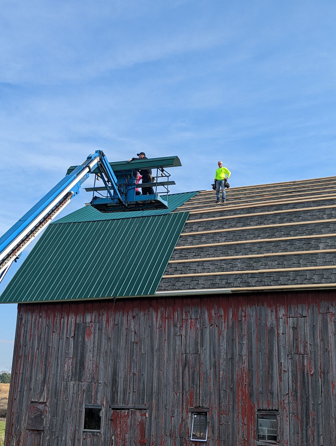 Barn roof being repaired with a lift; green metal and gray asphalt shingles; person working on the roof.