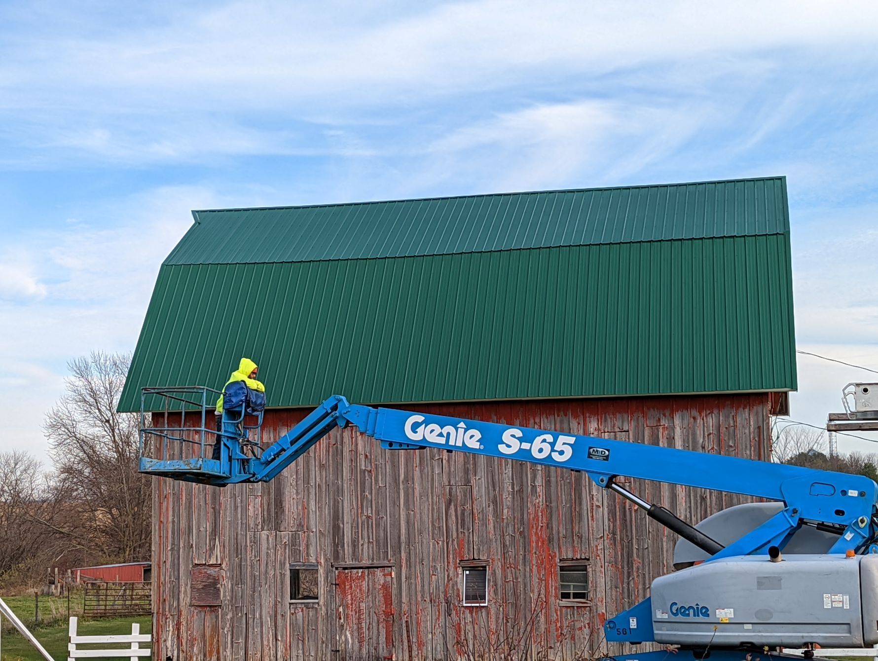 Person in a lift working on a green metal roof of a weathered red barn under a blue sky.