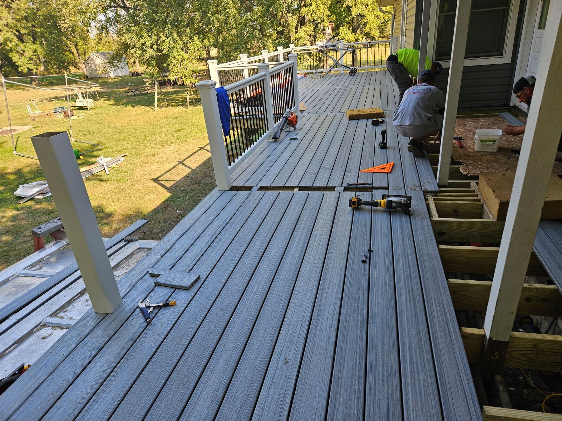 Workers installing gray composite decking on a porch. Tools and materials are scattered around the construction site.