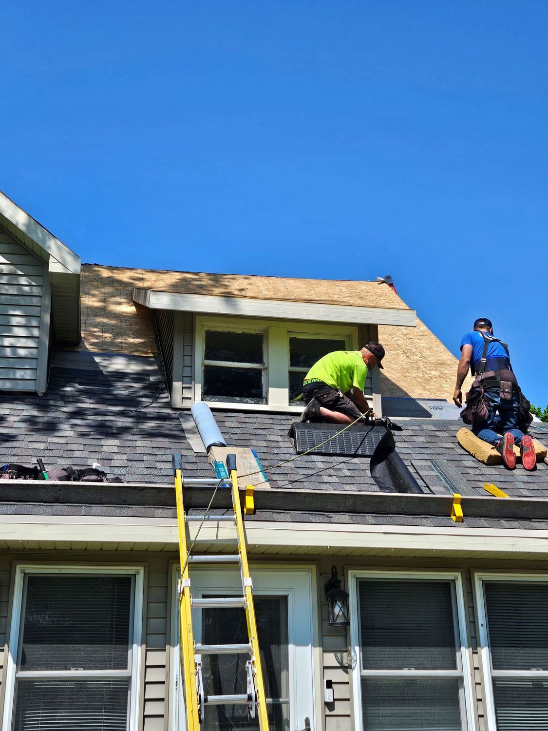 Roofers working on a house roof. One is kneeling, others are standing, blue sky.