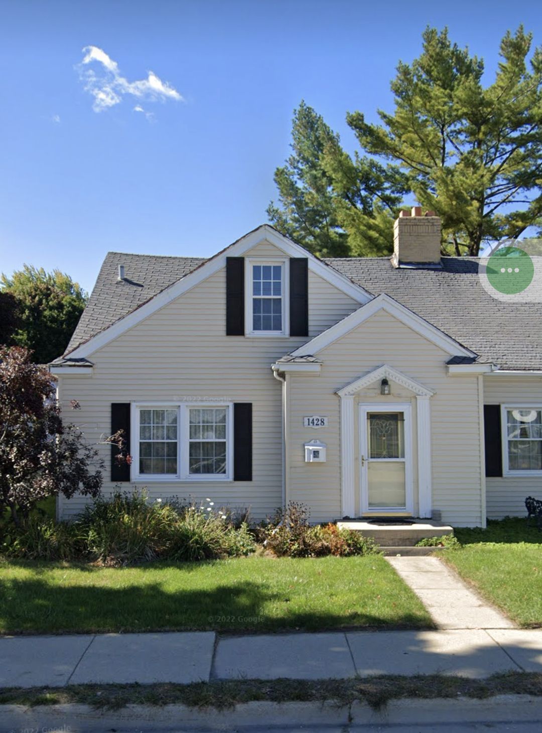 Cream-colored house with black shutters, small dormer window, and a concrete path leading to the front door.