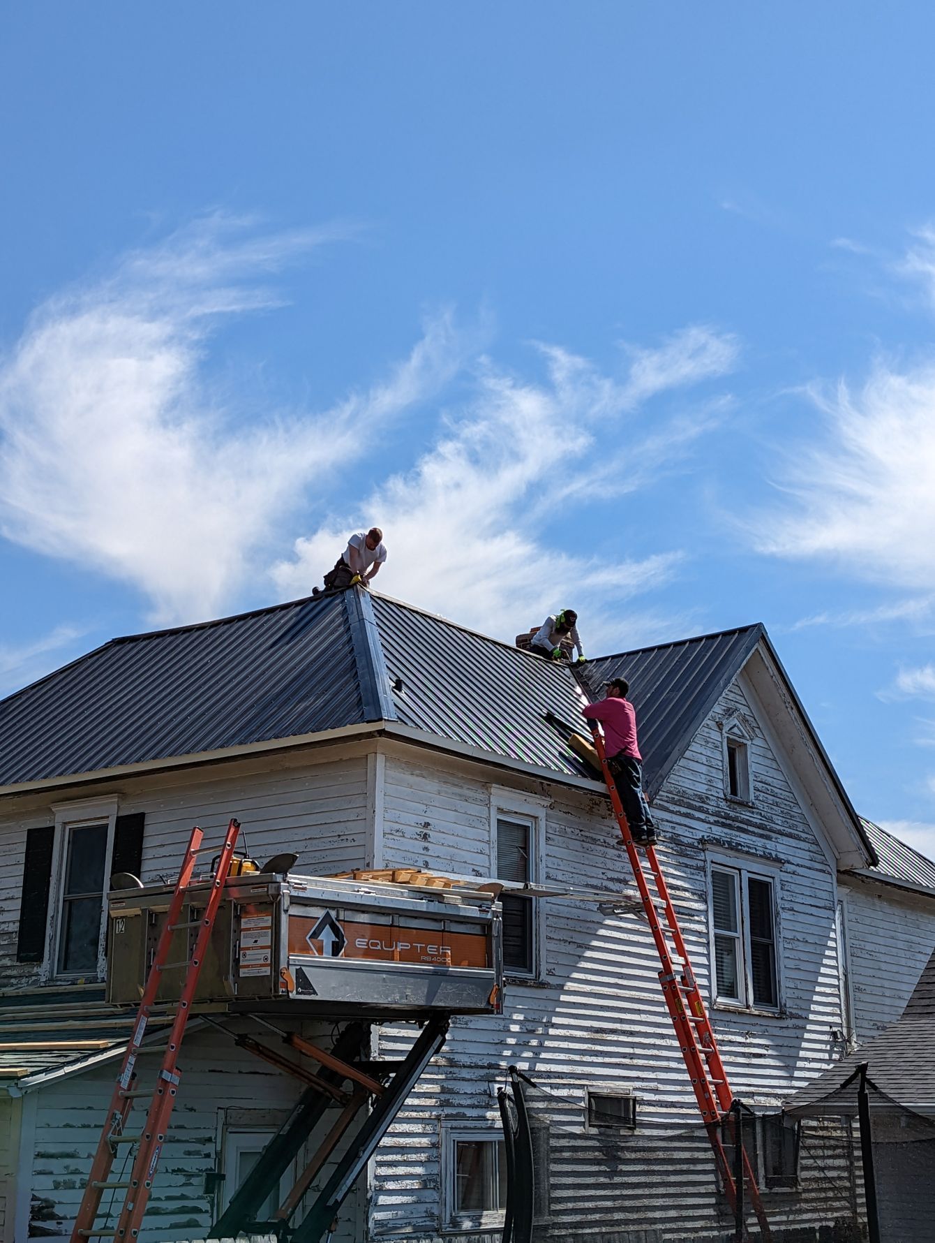 Roofers work on a metal roof, ascending ladders on a weathered white house under a blue sky.