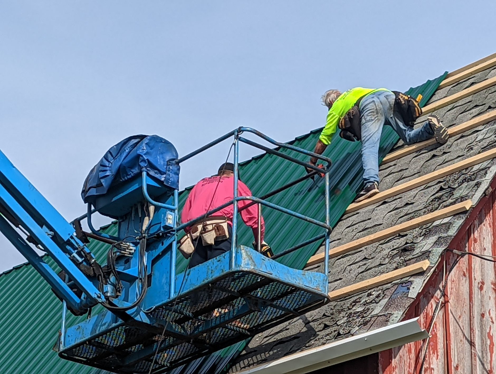 Two construction workers on a roof, one in a lift, repairing a metal roof. Blue sky in the background.