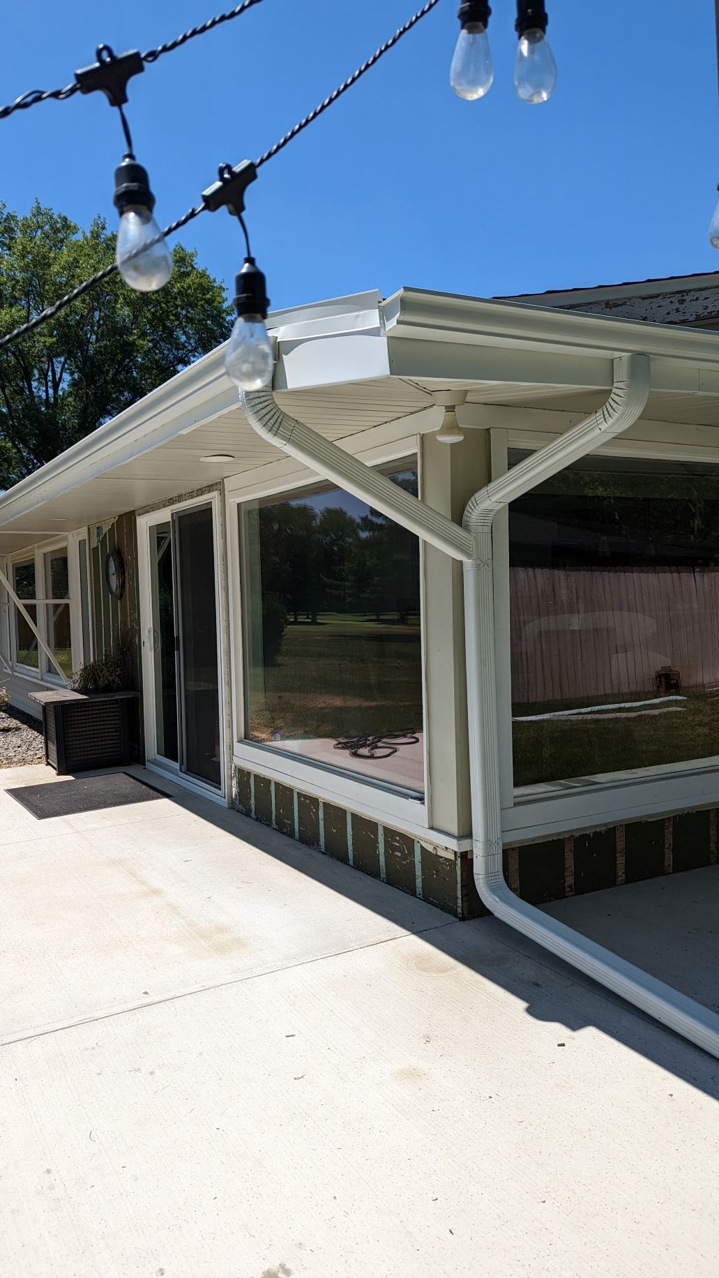 Exterior of a house with white gutters, large windows, and string lights on a sunny day.