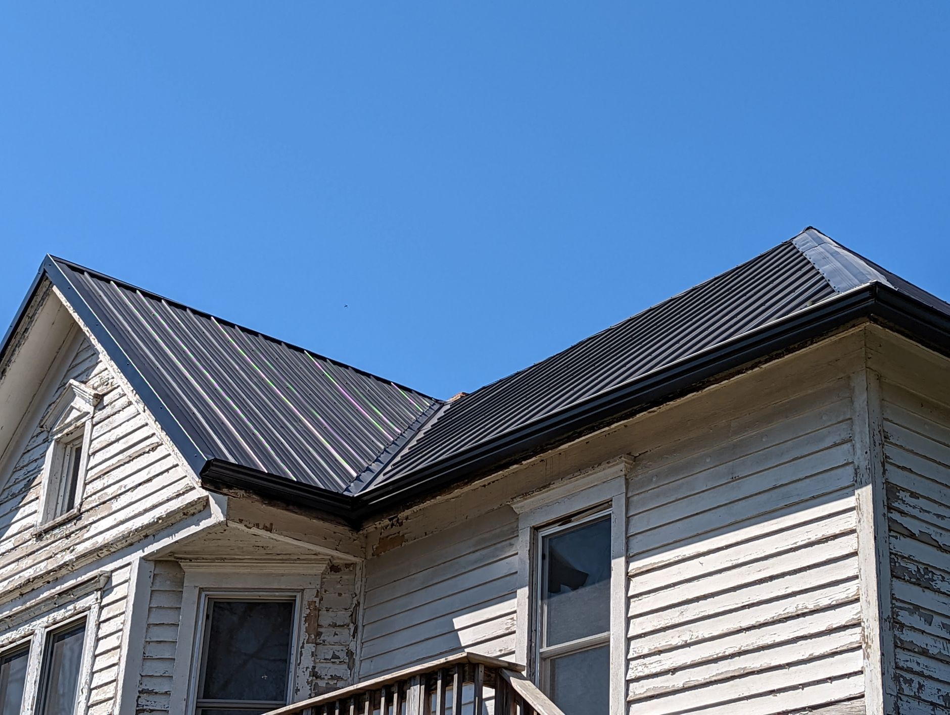 Weathered white house with a dark metal roof against a bright blue sky.