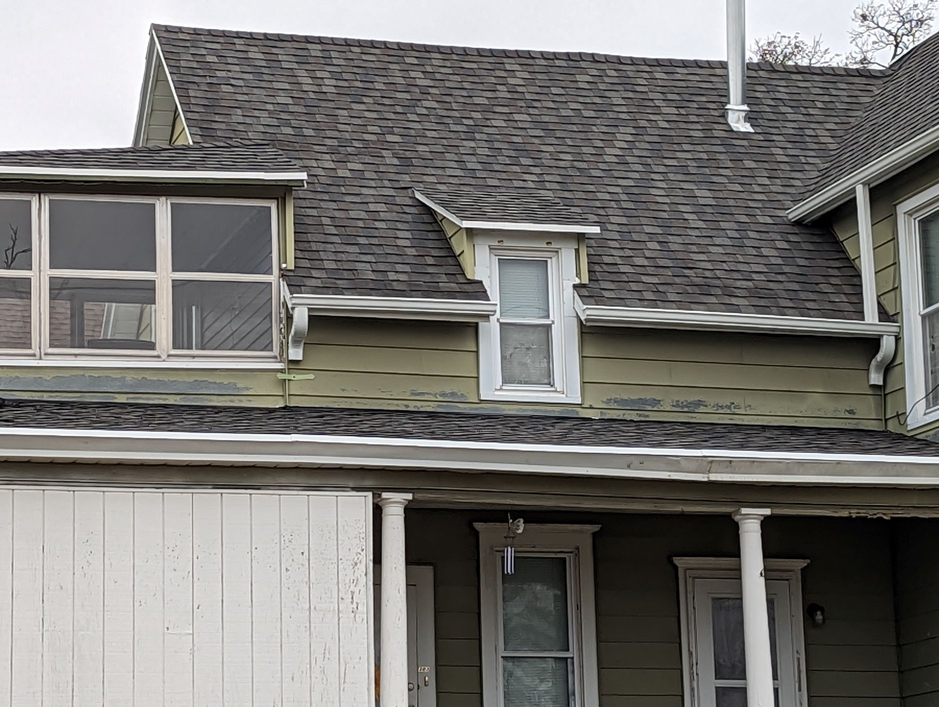 House with peeling green siding, dark roof, small dormer window, and porch with white pillars.