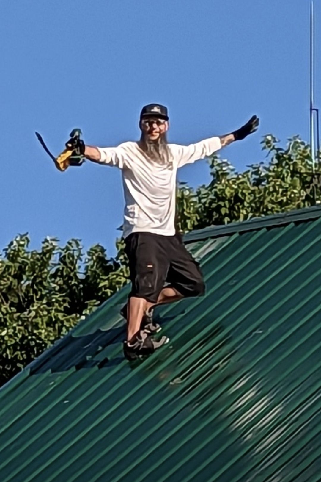 Man with tools on a green metal roof, arms outstretched, smiling; blue sky, trees.