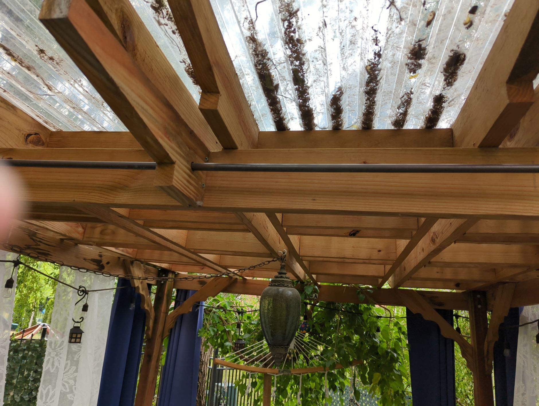 Wooden pergola with translucent roof panels, hanging lantern, and blue curtains.