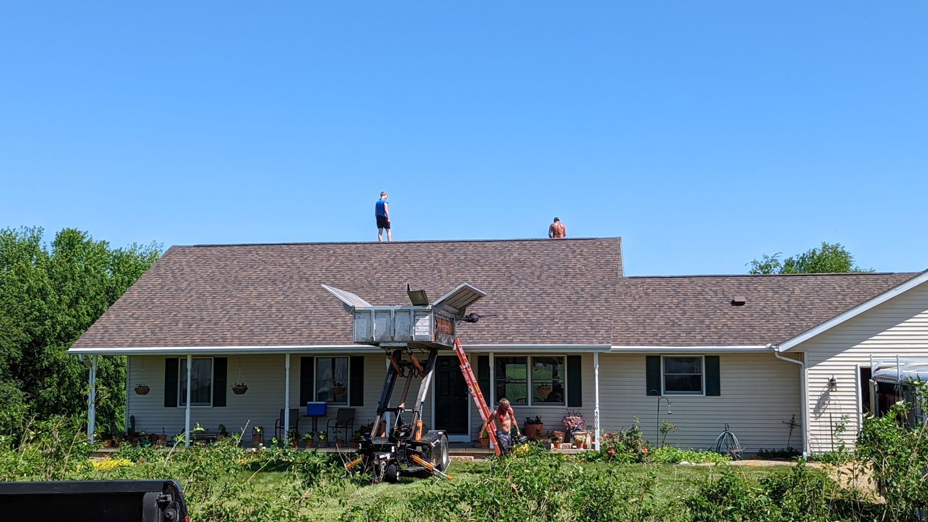 People on a roof, working. A tall ladder is leaned against the house. Sunny, blue sky.