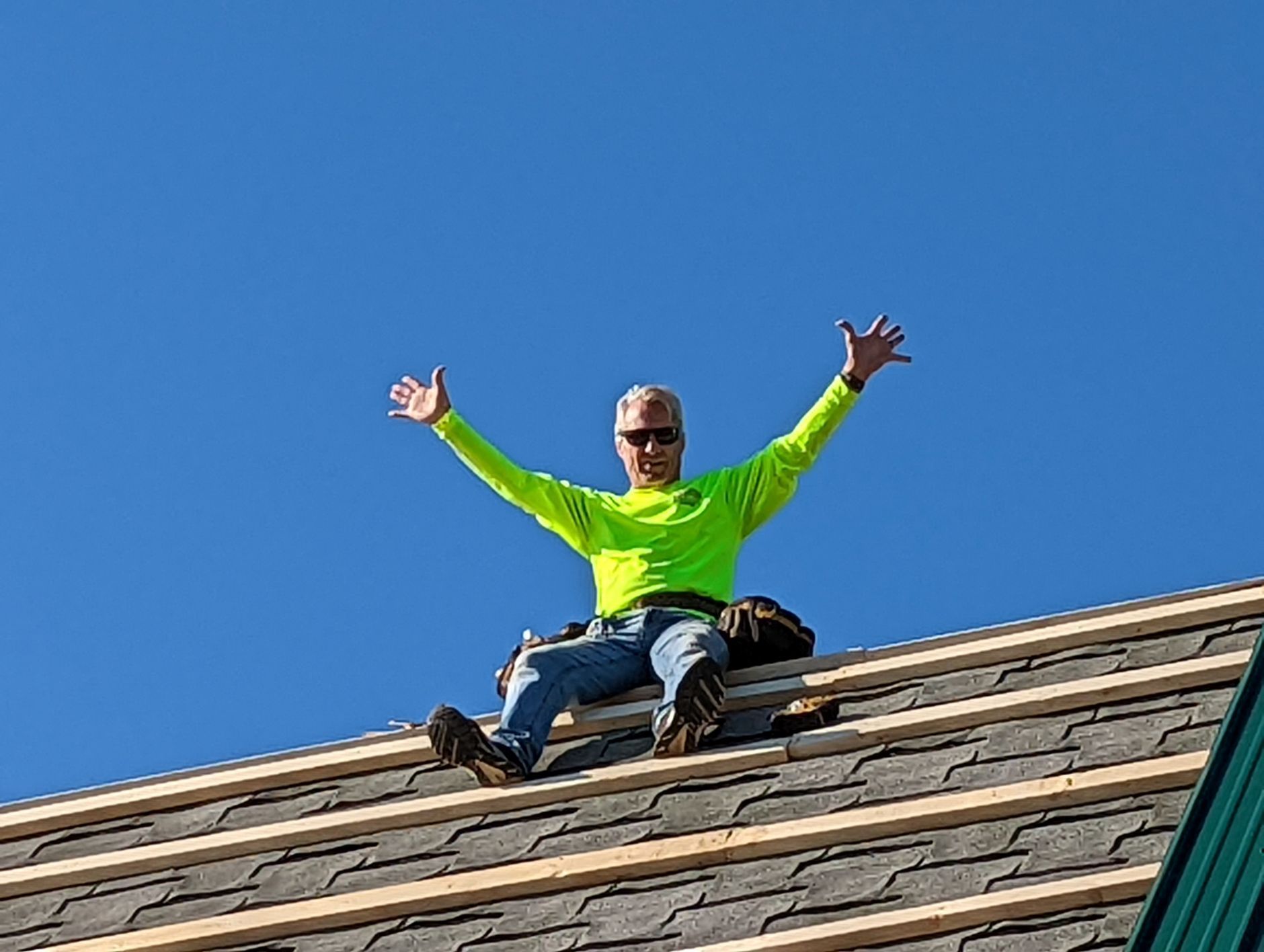 Man in neon shirt sits on rooftop, arms raised against blue sky.