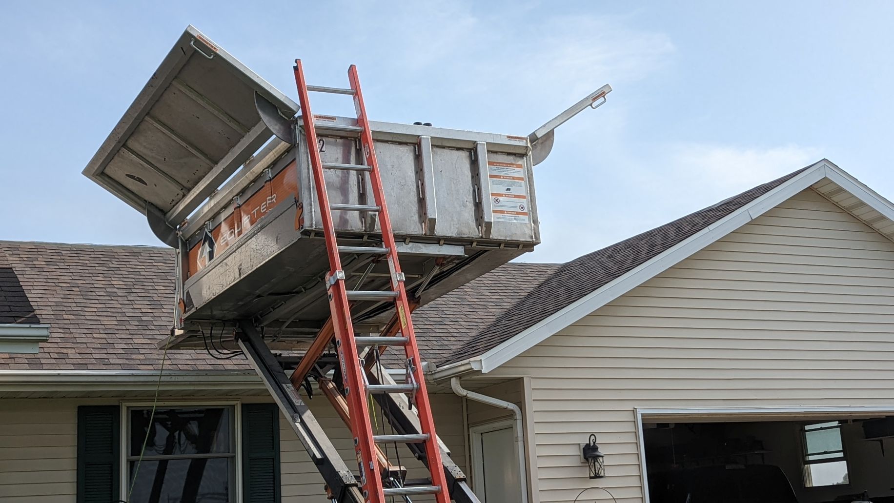 Elevated lift with ladder extending to a weathered roof. Beige house with brown shingles. Sky is clear and blue.