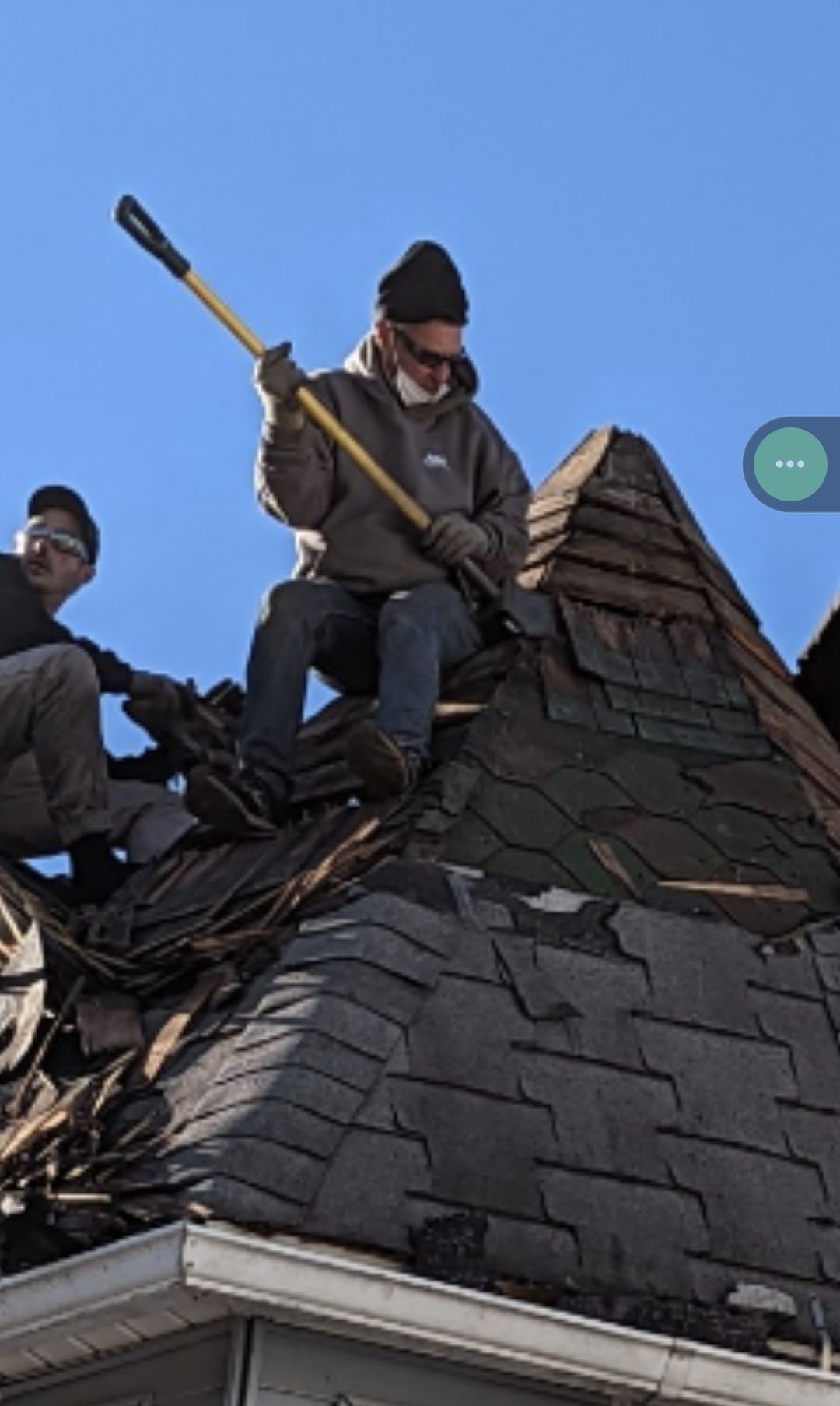Two workers removing shingles from a rooftop under a blue sky; one uses a tool, the other assists.
