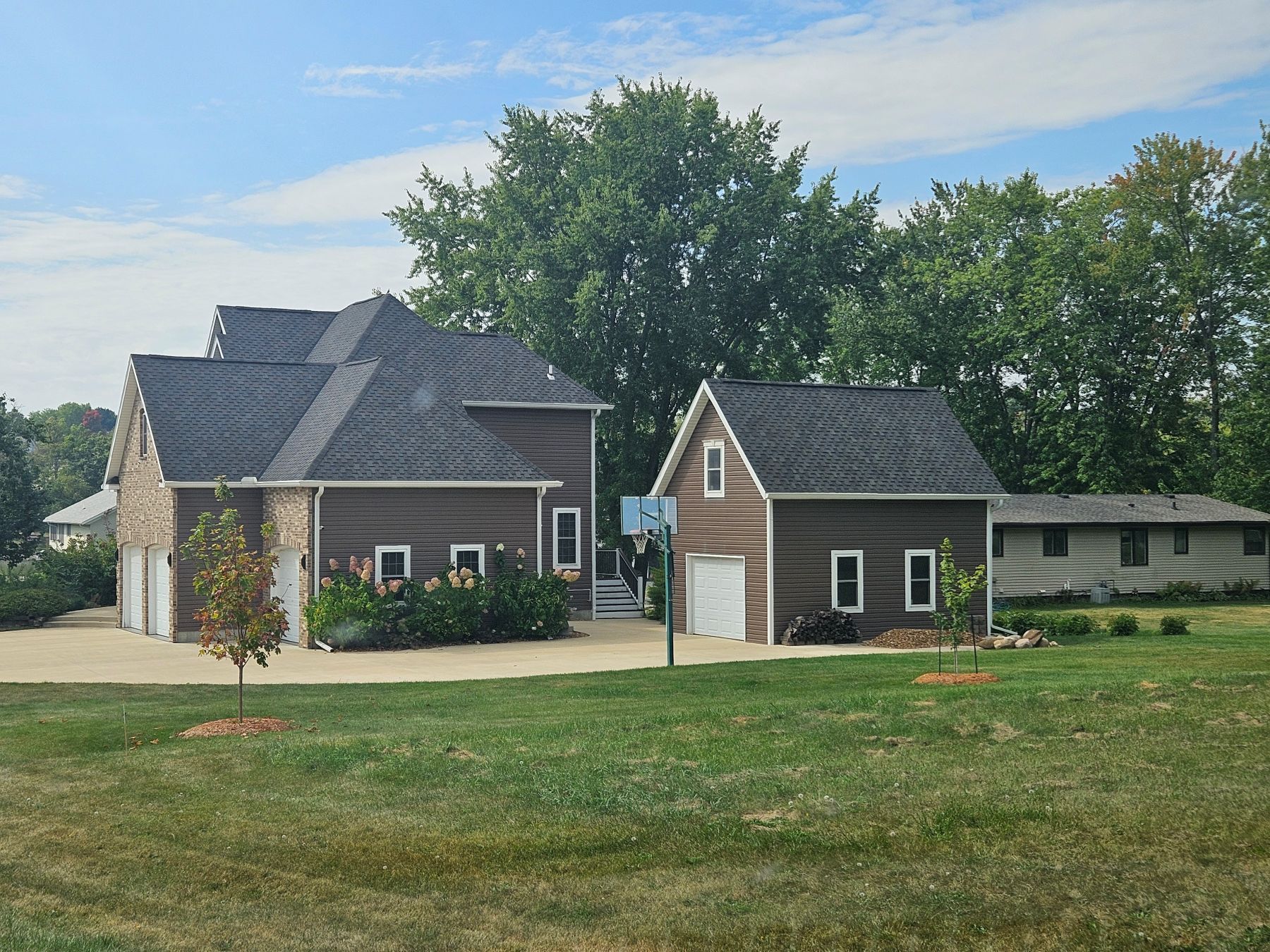 Brown-sided house and detached garage with dark roof, basketball hoop, and green yard under a blue sky.