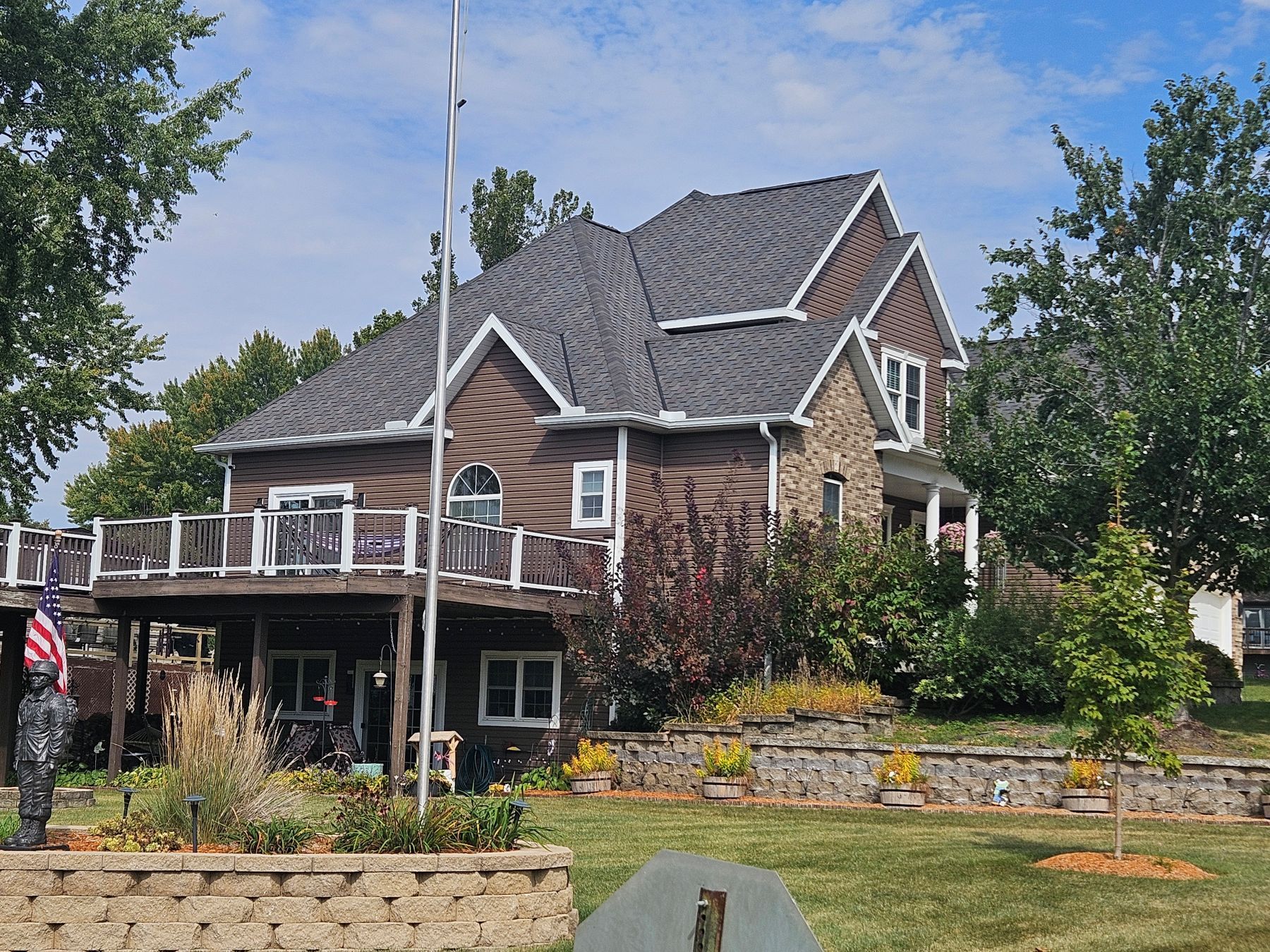 Two-story house with brown siding, stone accents, and a deck, under a blue sky, on a grassy lawn with trees.