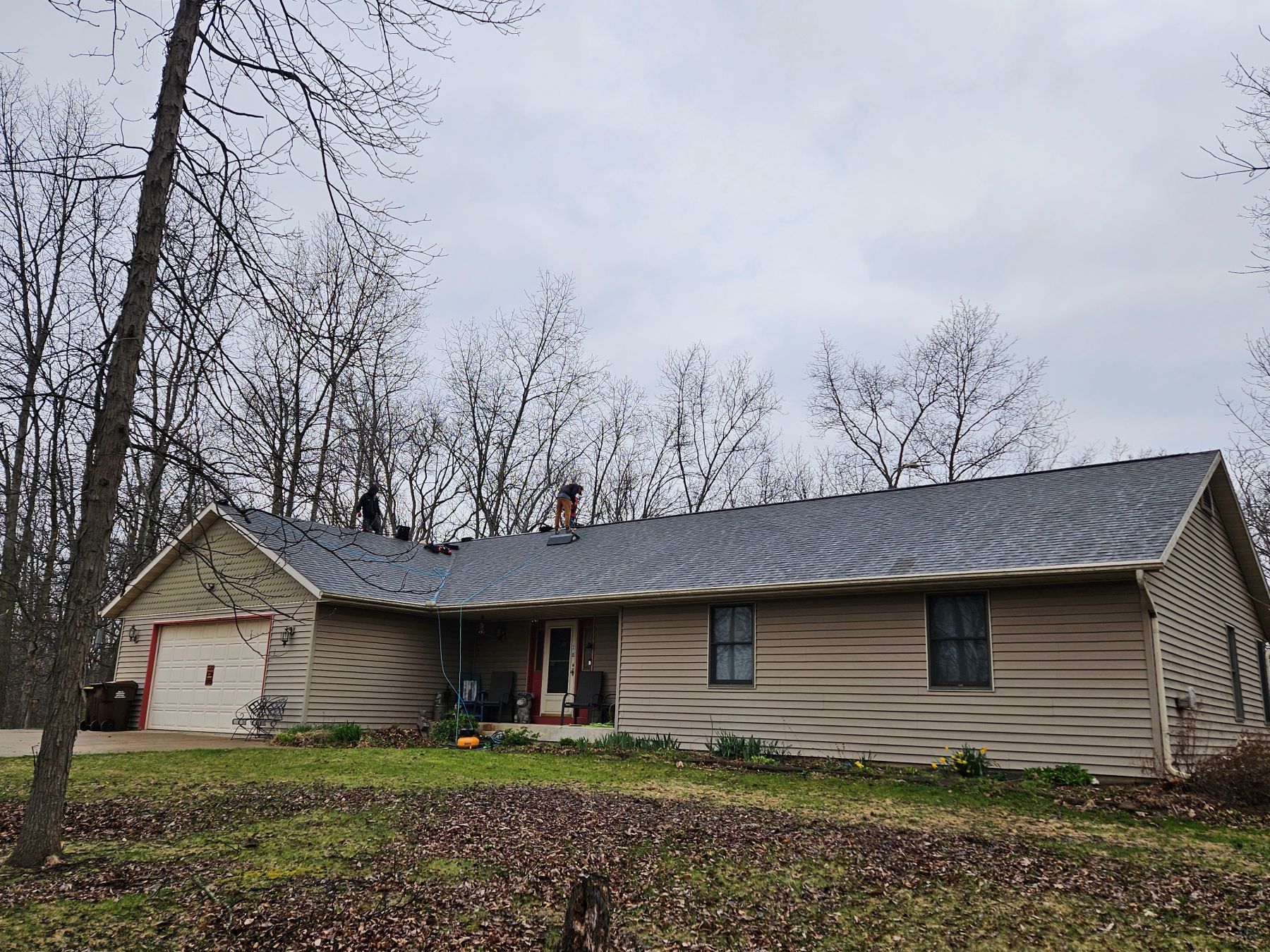 House with a new gray roof, beige siding, and surrounding bare trees under a cloudy sky.