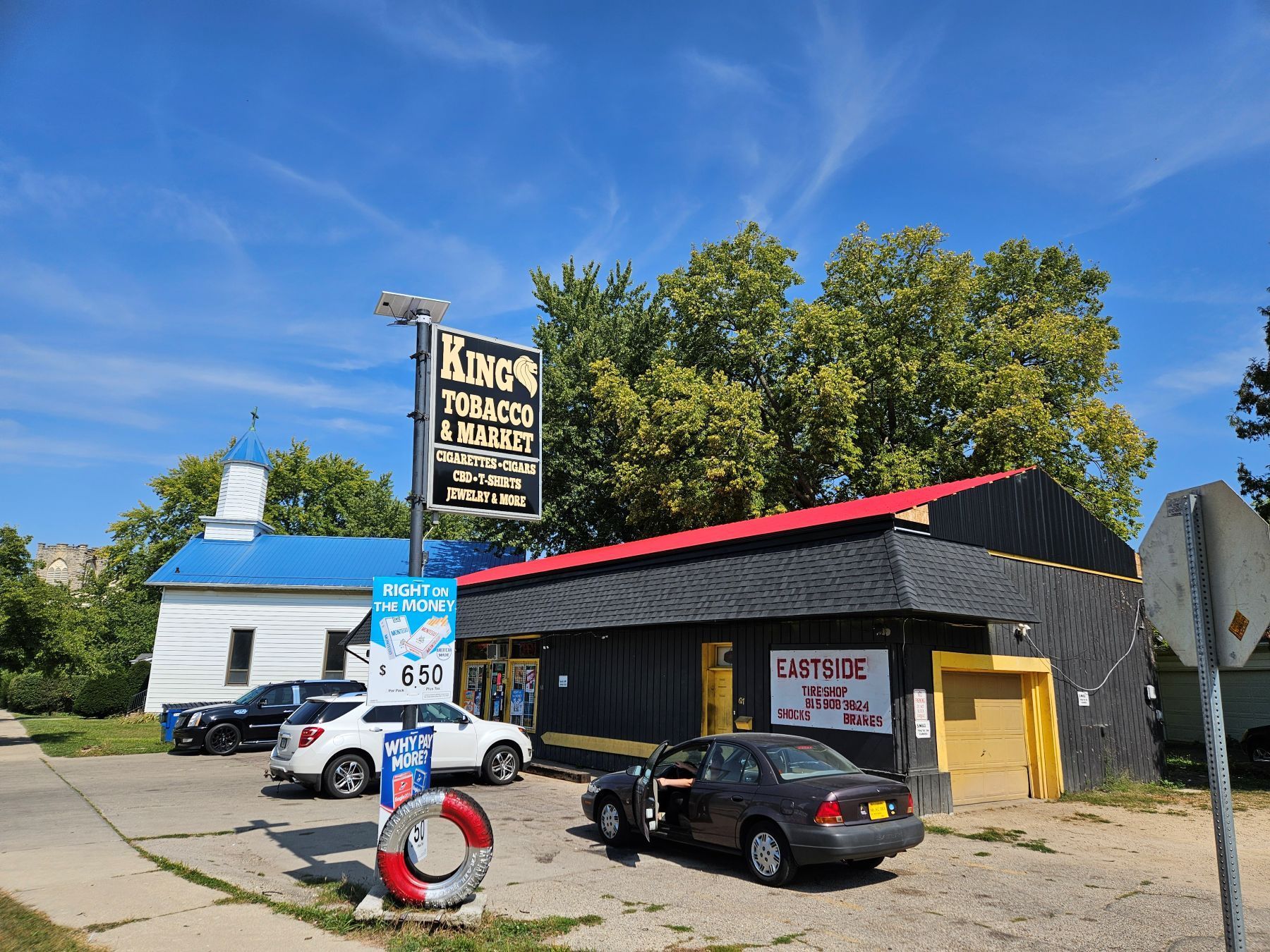 Kino Tobacco & More store with black facade, red roof, blue sky, cars, and a small white building next door.