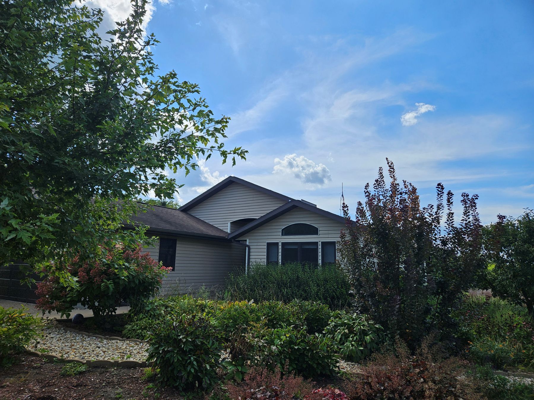 House partially hidden by green and red bushes under a blue sky with clouds.
