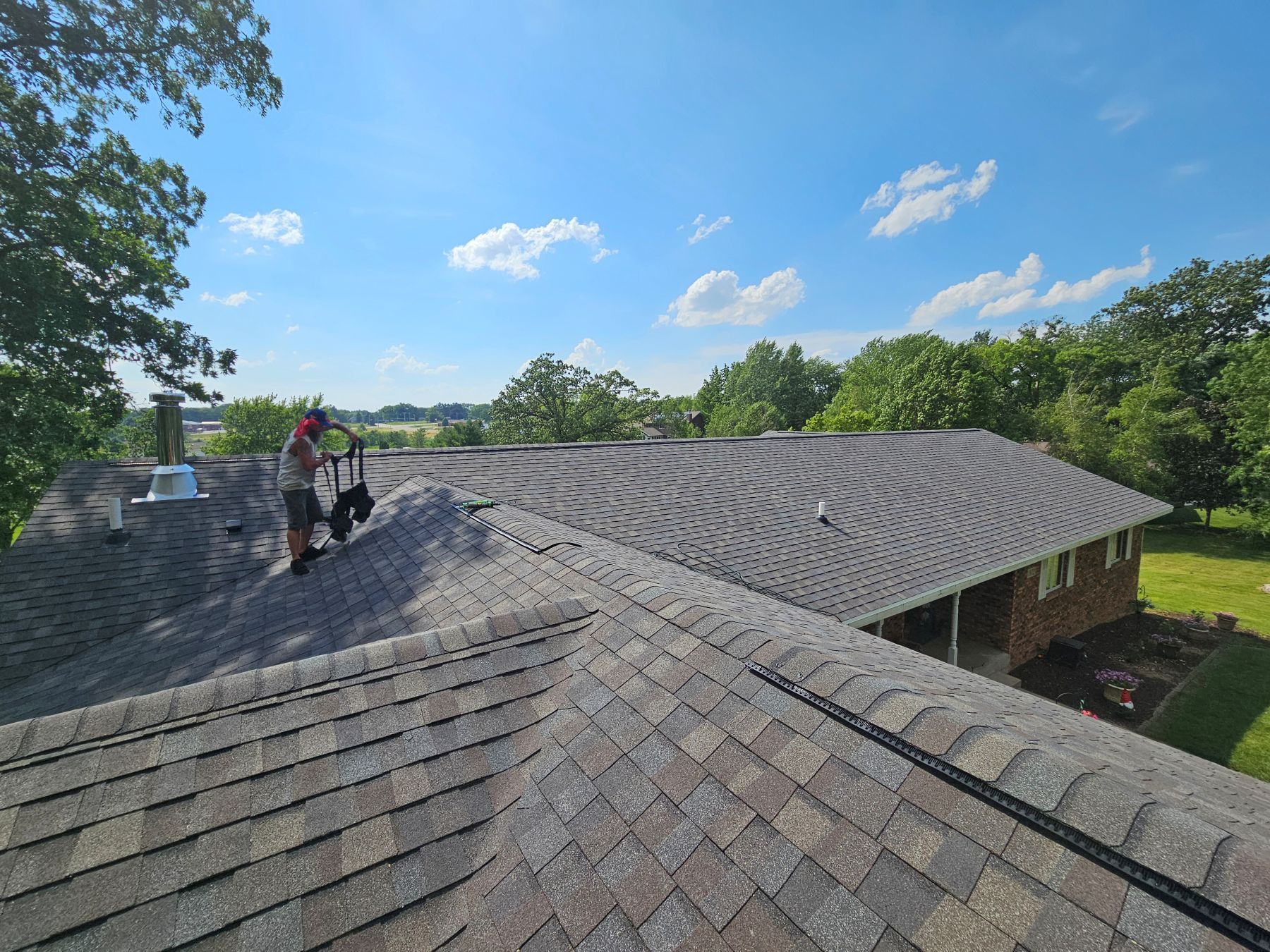 Person on a roof, inspecting shingles on a sunny day. Green trees and blue sky in the background.