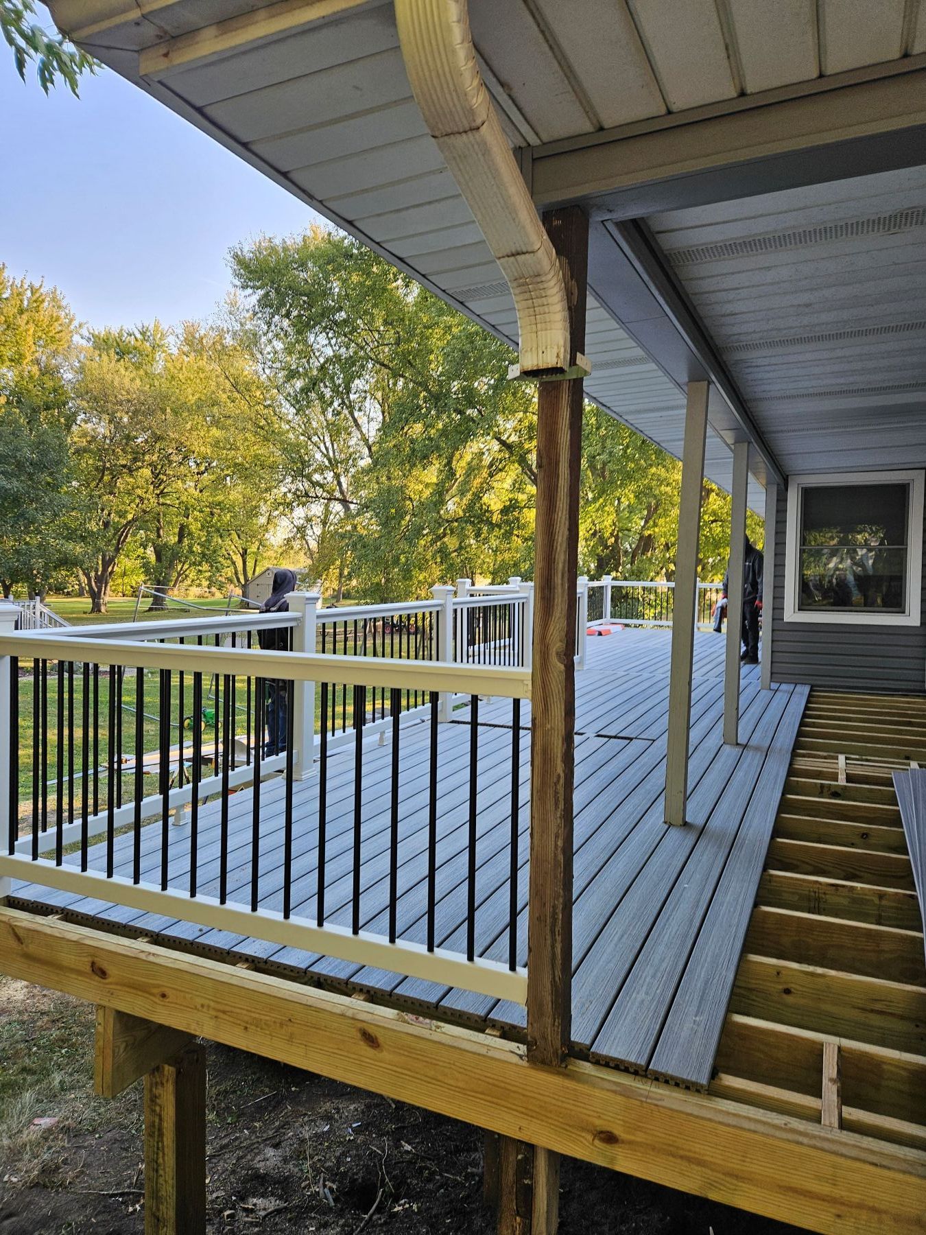 Partially constructed gray deck with black railing and a brown support post, trees in the background.