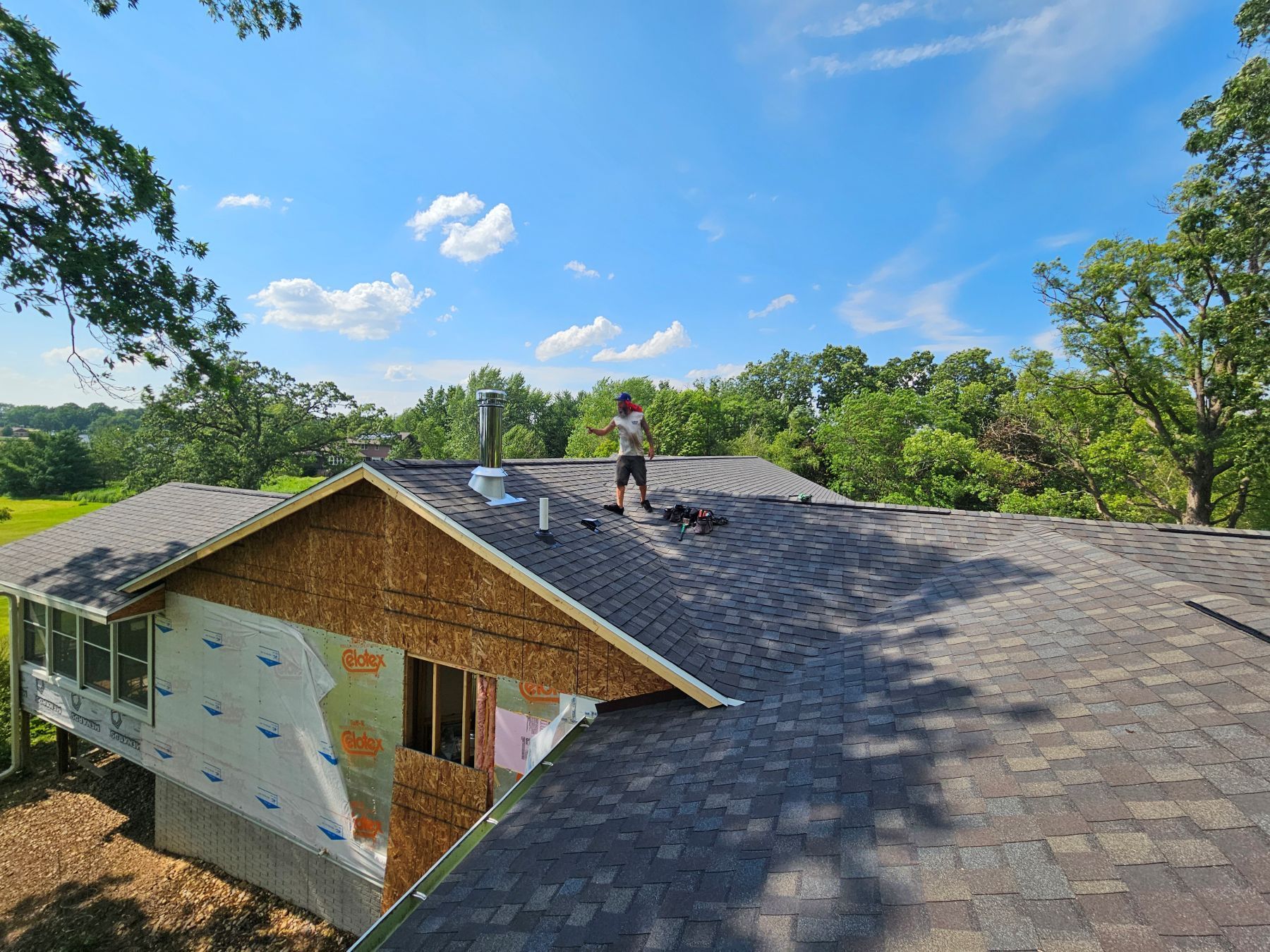 Roofer working on a house roof. Blue sky with clouds. Trees in the background.