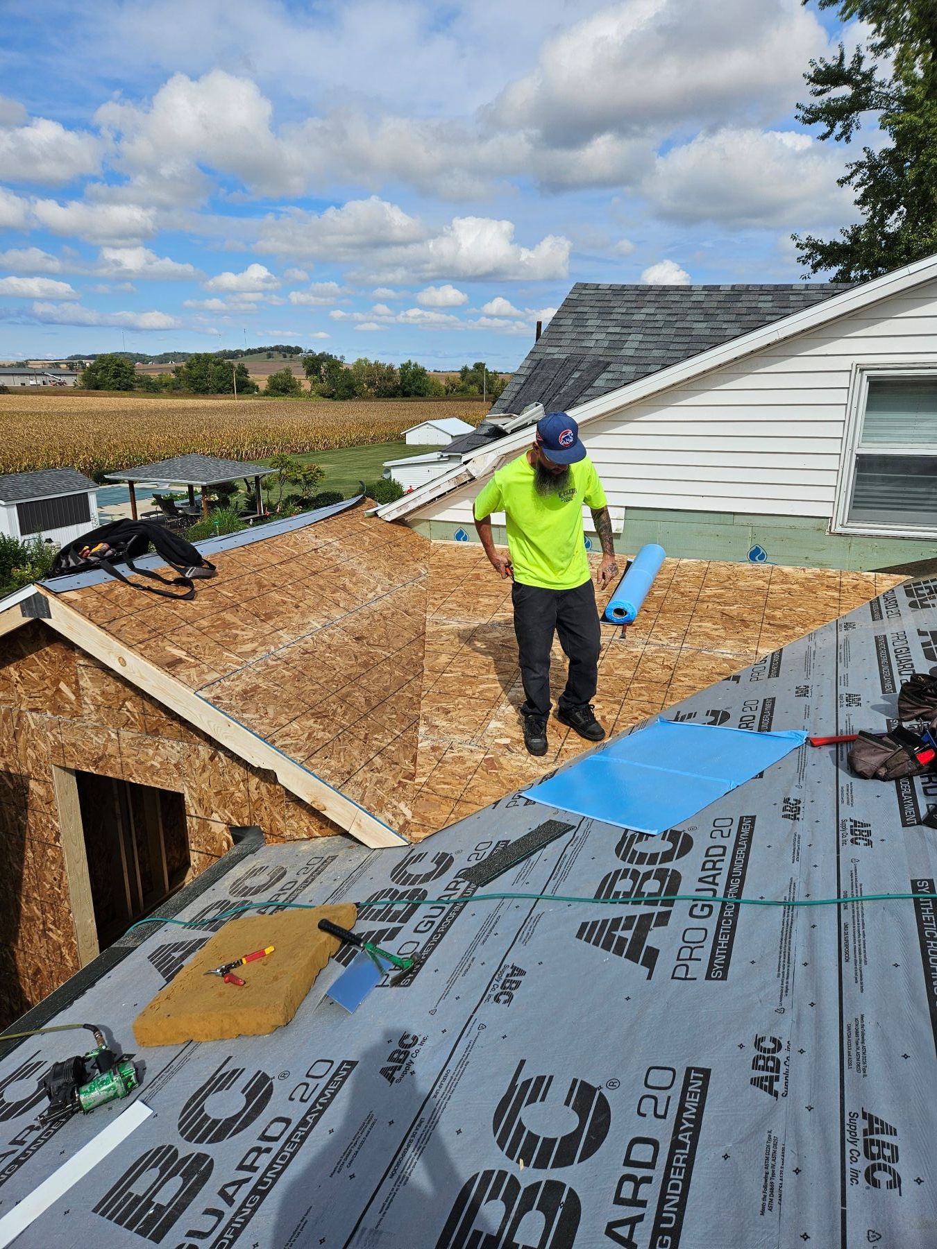 Roofer on a partially built roof, with blue underlayment and plywood visible, outdoors on a sunny day.