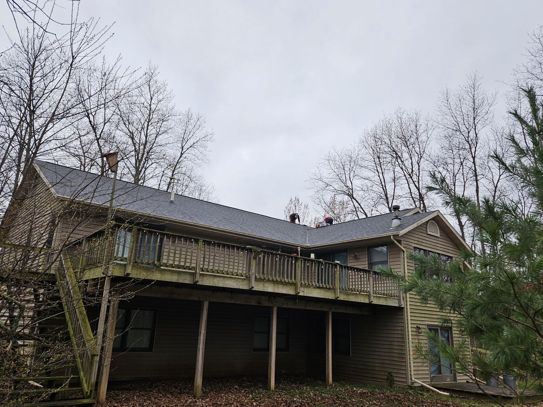 Two-story house with a large wooden deck. Gray roof, tan siding, surrounded by bare trees under a cloudy sky.