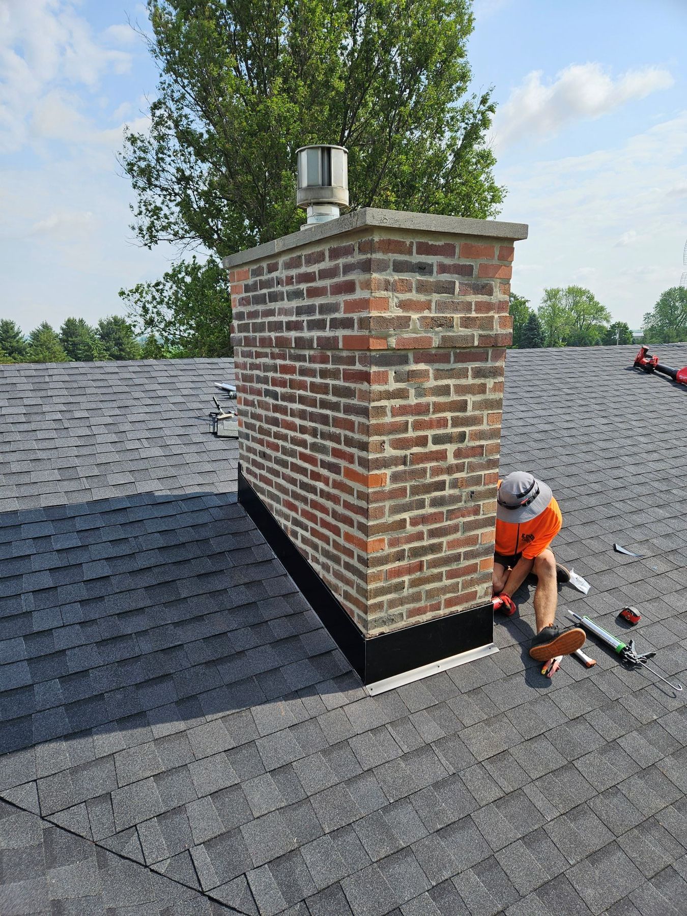 Roofer installing flashing around a brick chimney on a shingled roof under a blue sky.