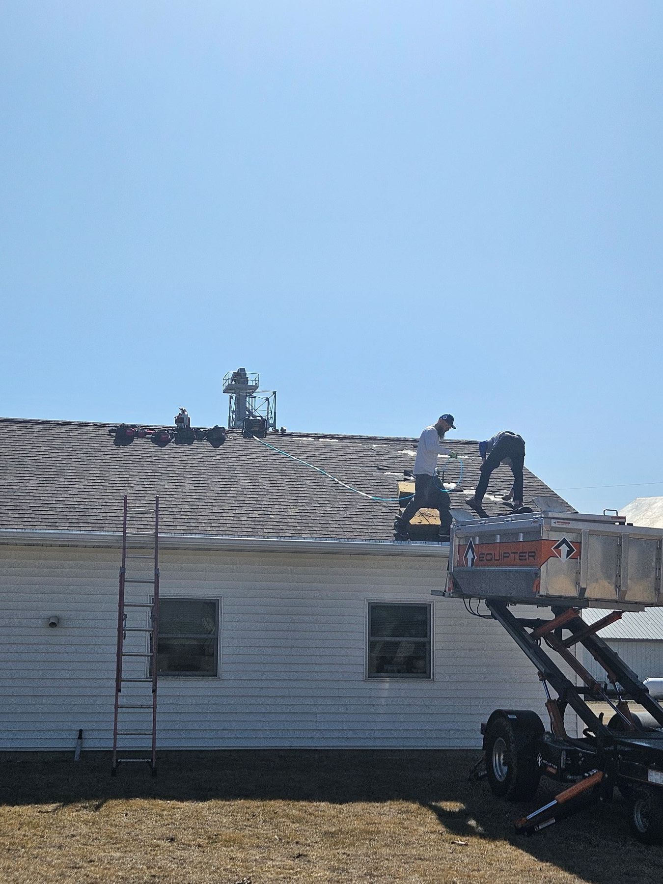 Two workers on a roof, one in a lift, repairing shingles on a white building under a blue sky.