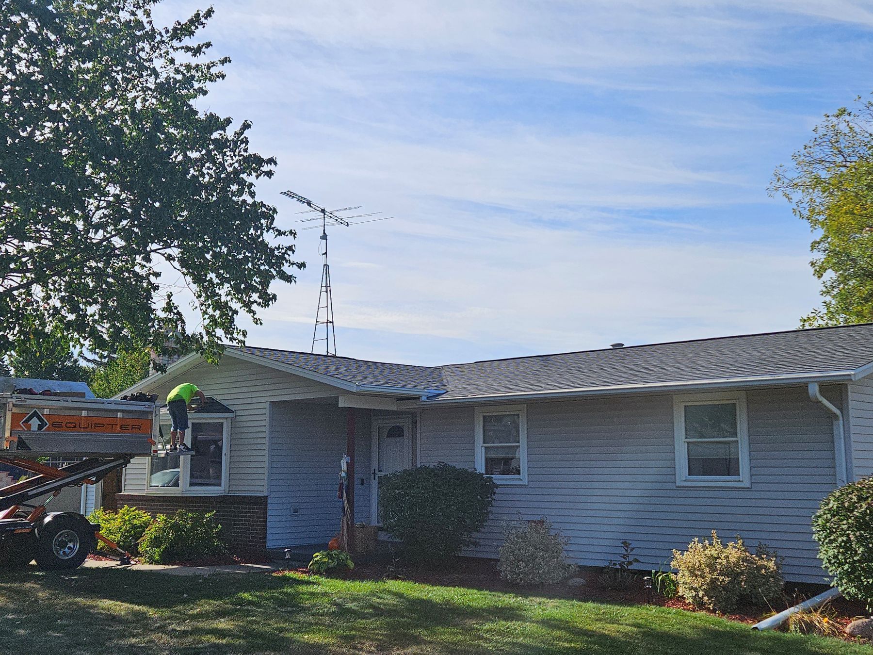 Person on a lift working on the roof of a gray, one-story house on a sunny day.