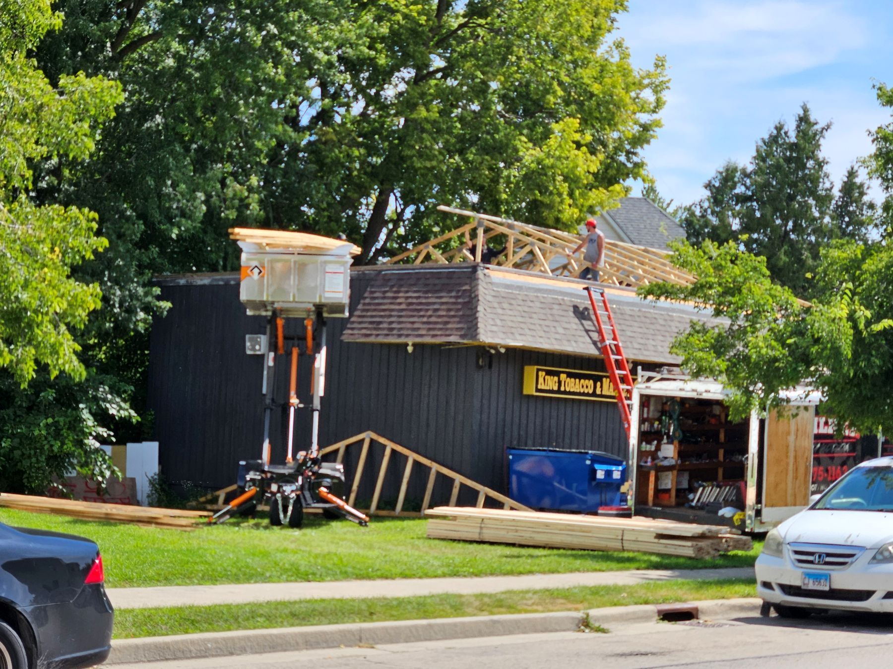 Construction on a building roof. A worker on a ladder, trusses, and a lift are visible. Green trees and a car are in the setting.