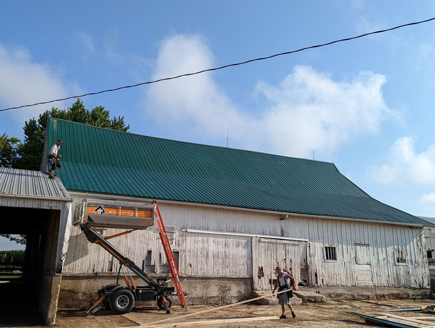 Barn with green metal roof, people working, ladder, lift, blue sky.