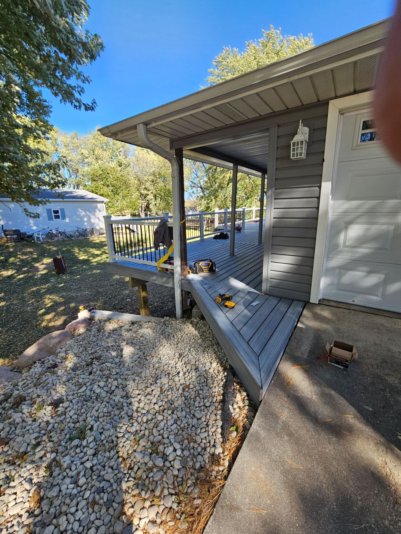 Grey deck and ramp leading to a house with gray siding. Sunlight, trees, and gravel ground visible.