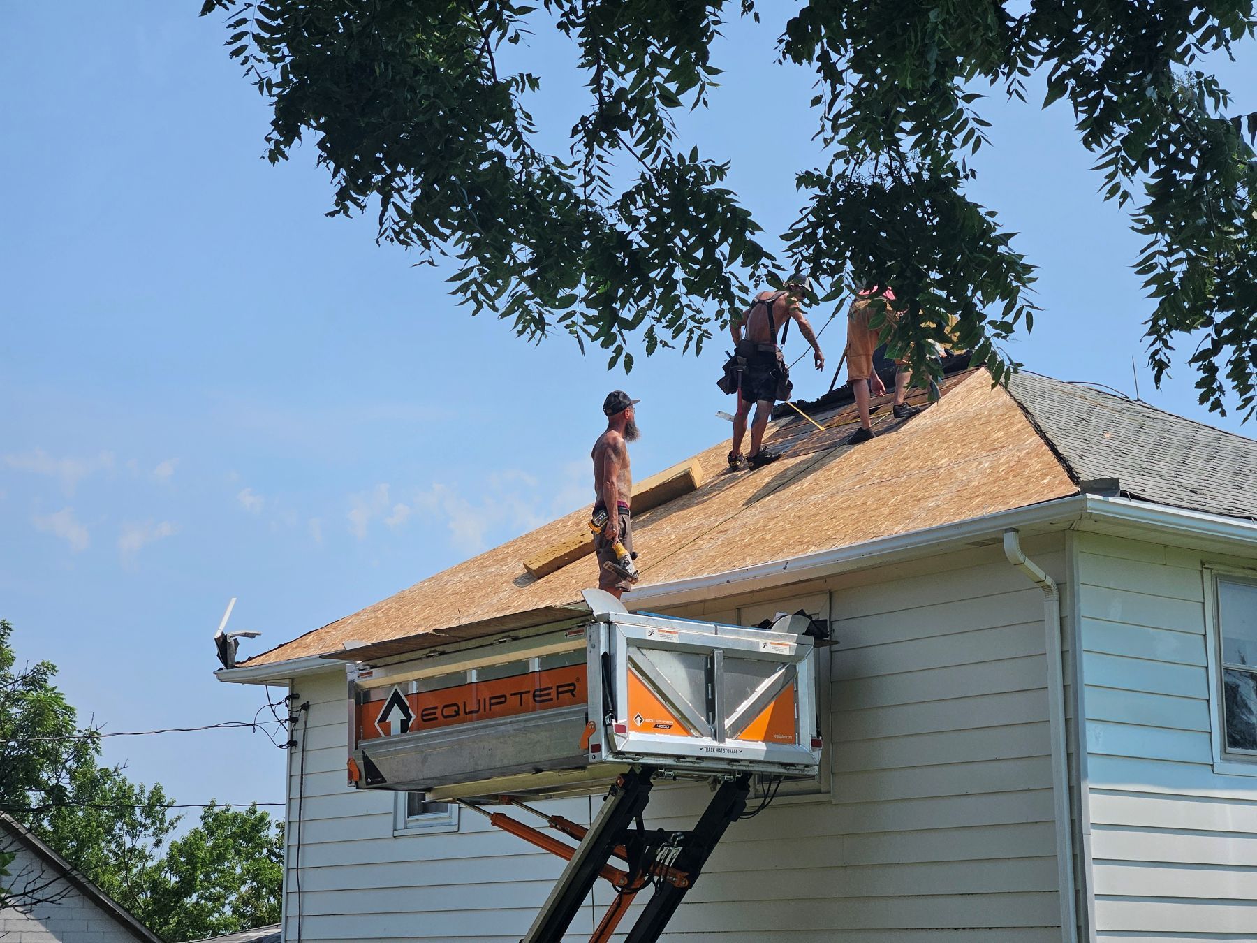 Men roofing a house, working on shingles. A lift provides access. Blue sky.