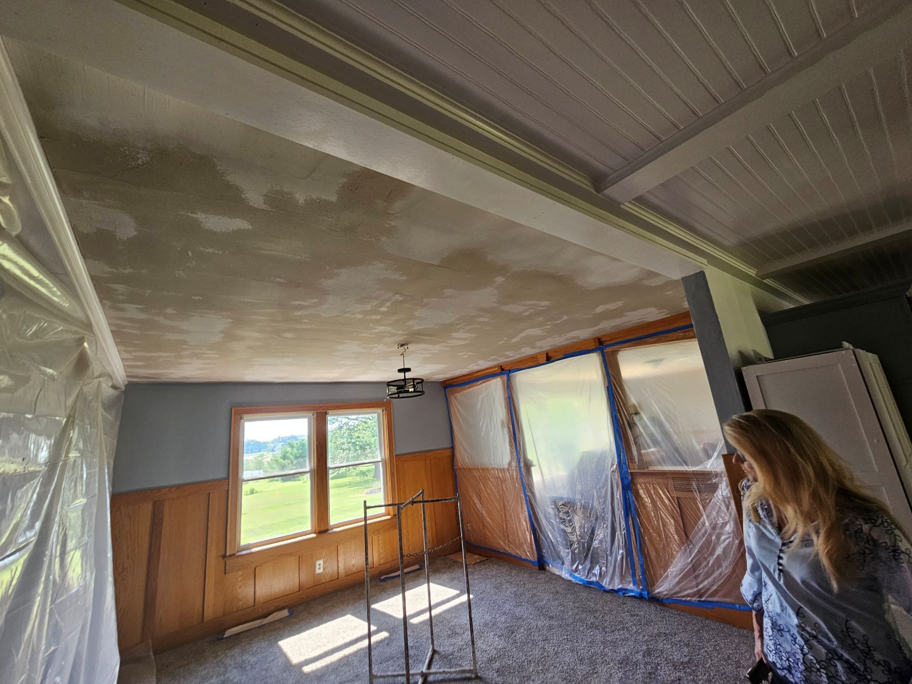 Room with plastic sheeting, wood paneling, a woman looking on, and a partially refinished ceiling.