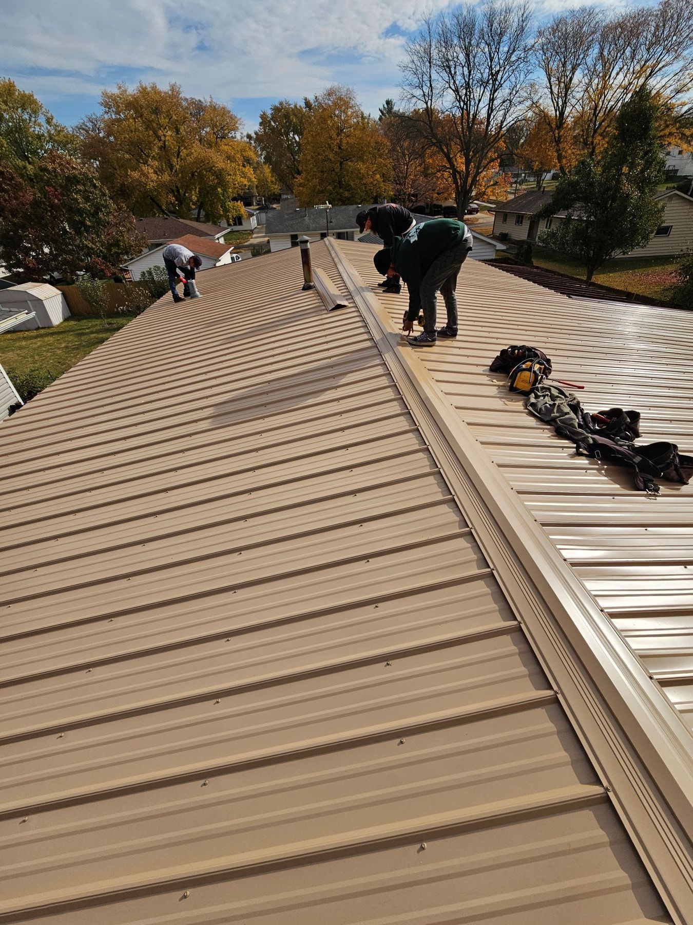 Three workers installing a tan metal roof on a residential building on a sunny day.