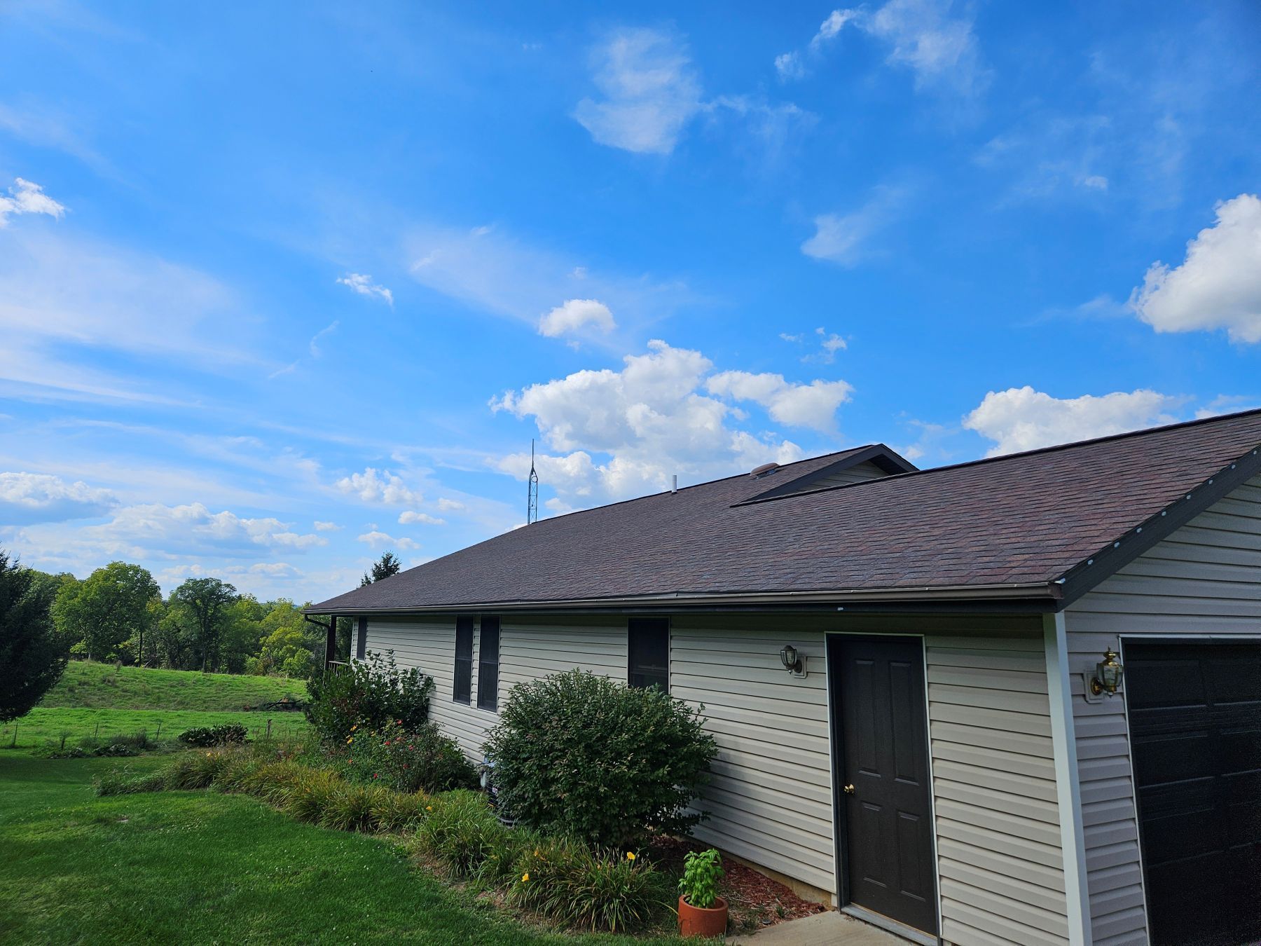 A house with brown roof and light siding against a blue sky with clouds; green lawn and foliage in the foreground.
