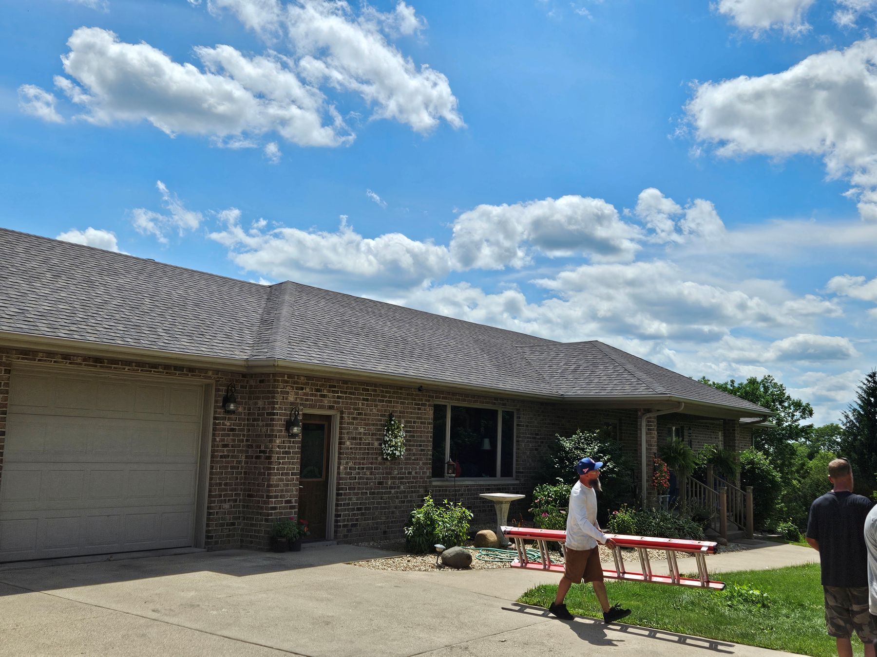 House with brown brick facade, garage, and blue sky. People walk on the driveway.