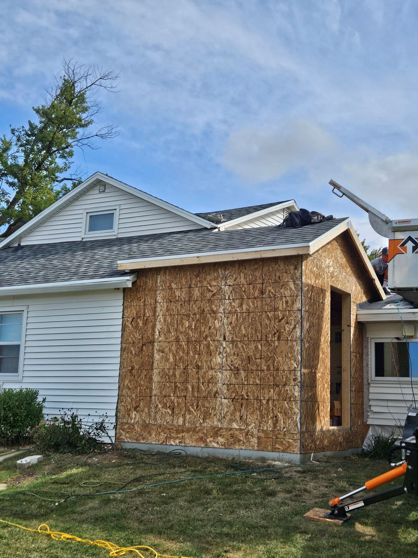 Exterior house renovation in progress; new addition framed with OSB siding; blue sky.