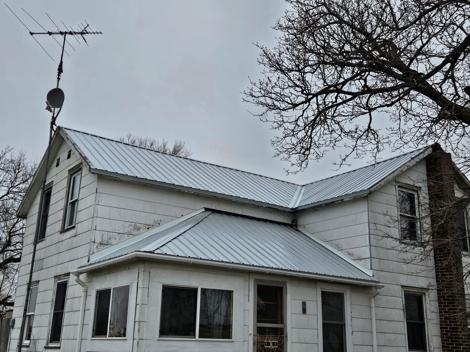 White farmhouse with a metal roof, antenna, and chimney against a cloudy sky.